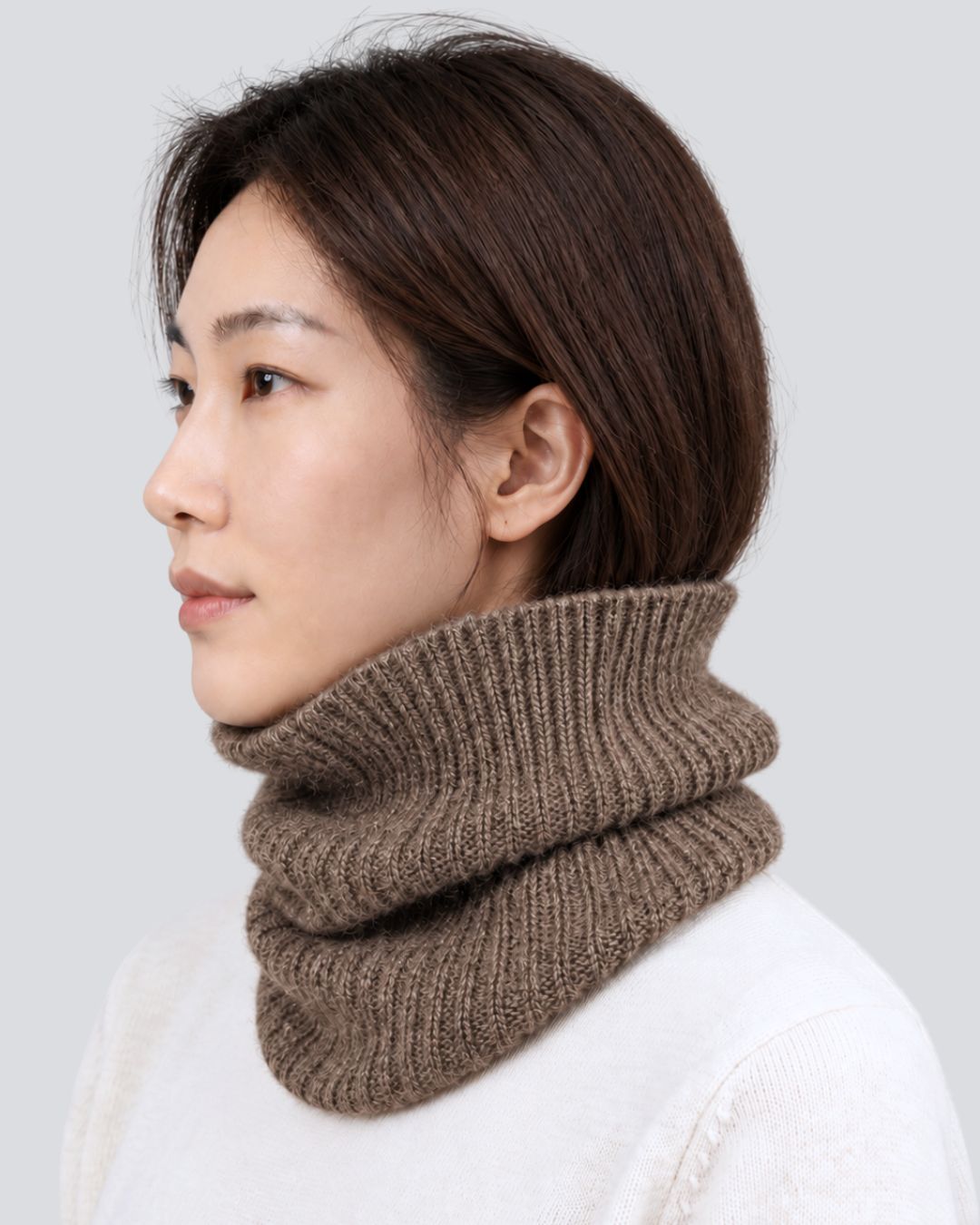 A woman shown in profile wearing a thick, ribbed brown knit cowl neck warmer over a white sweater, against a light gray studio background.