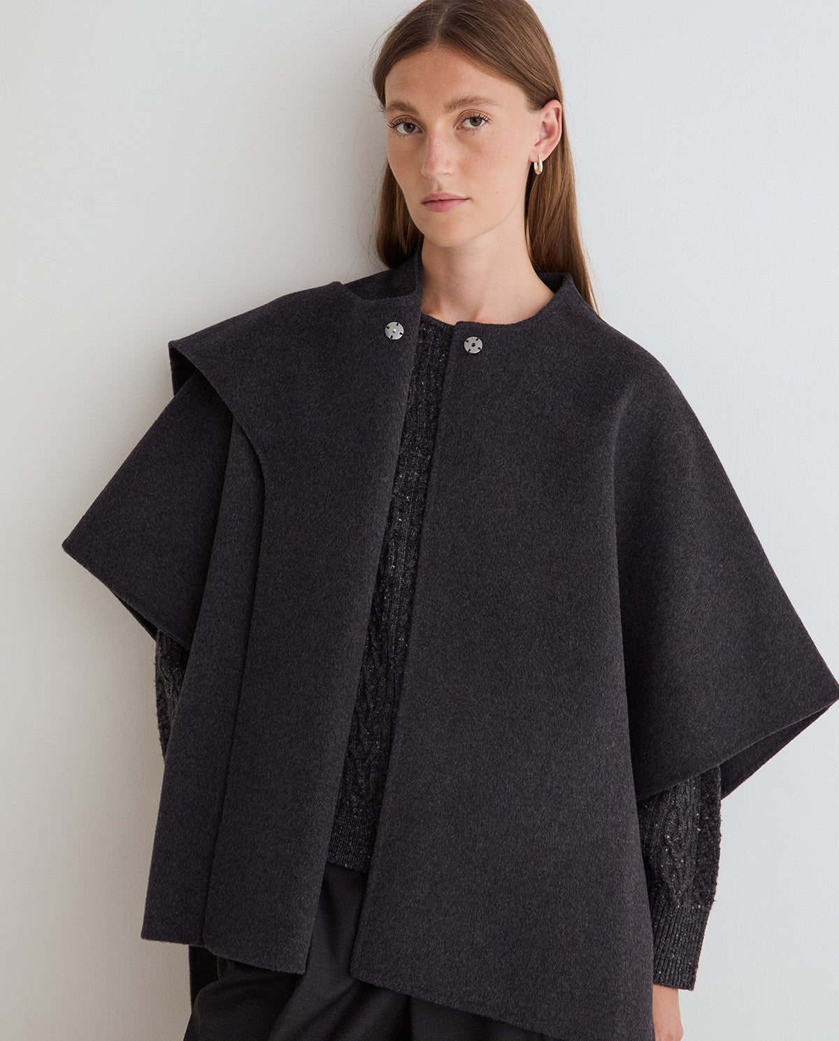 A woman wearing a dark charcoal gray cape-style coat with wide draped sleeves, a round neckline, and silver-tone decorative buttons, photographed against a plain light gray wall.