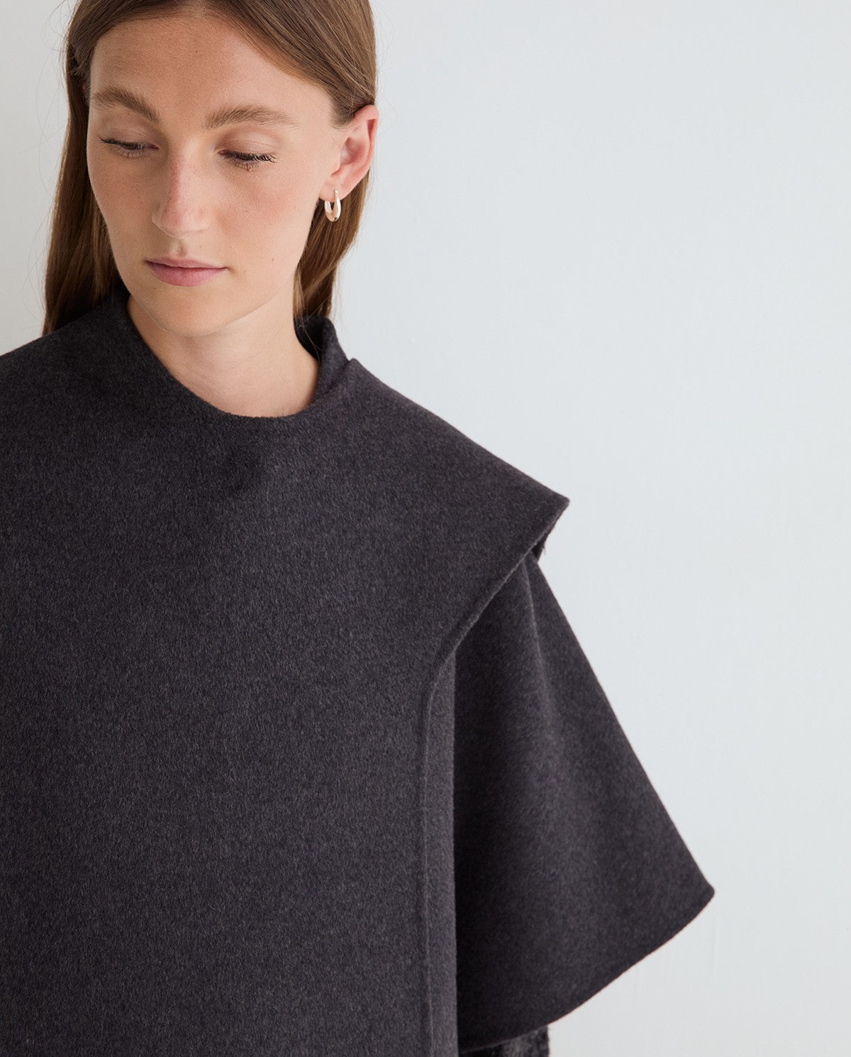 A close-up of a model wearing a dark charcoal gray cape-style sweater with a high mock neck and wide draped sleeves against a plain light gray background.