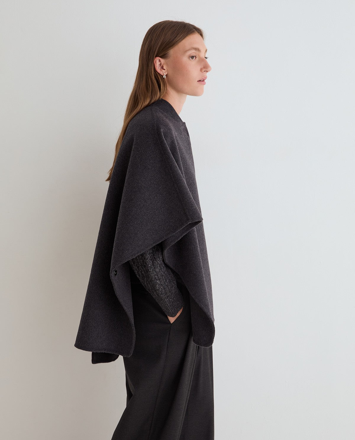 A woman in side profile wearing a dark charcoal gray draped poncho over a textured black top and black trousers, standing against a plain light gray studio background.