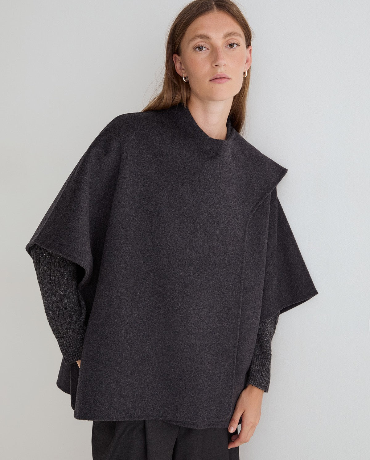 A woman wearing a dark charcoal gray cape-style knit top with textured knit sleeves, photographed against a plain light gray studio background.
