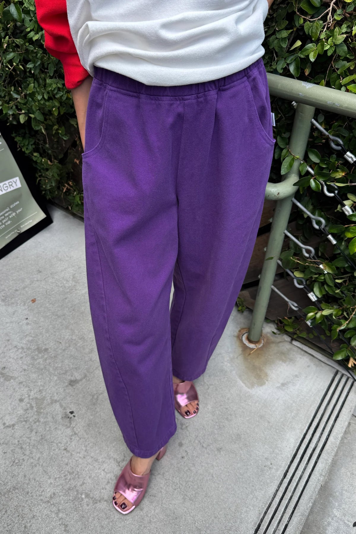 Eggplant purple Arc Pants, front view with hands in pockets, styled with a white and red top and pink sandals