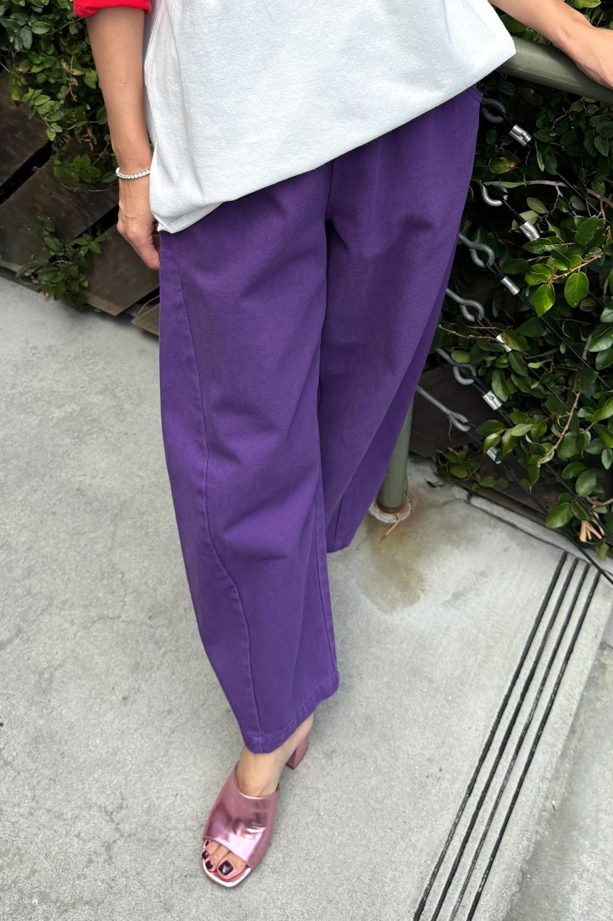 Eggplant purple Arc Pants, angled front view showing rounded legs and cropped hems, worn with pink sandals.
