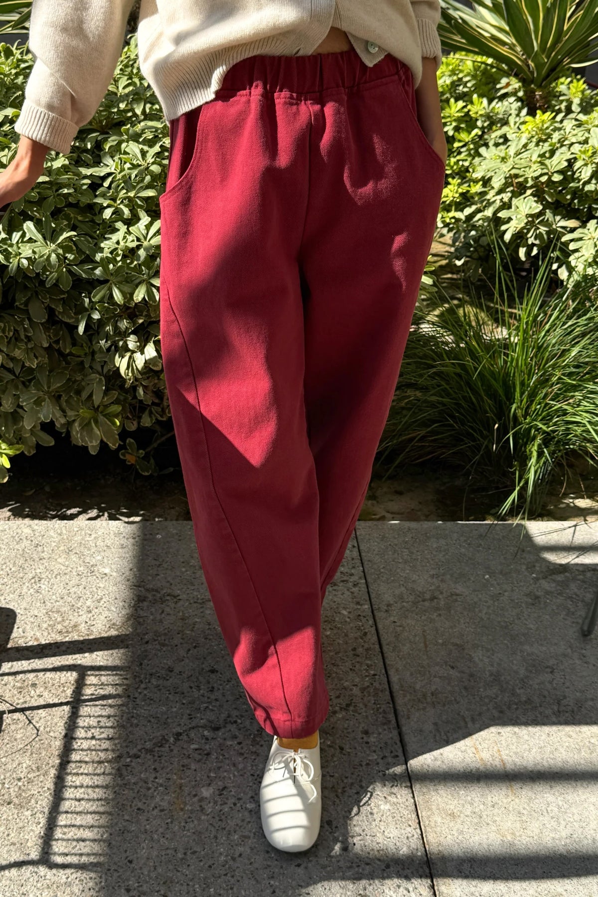 Oxblood Arc Pants, front view showing elastic waist and curved legs, worn with a cream cardigan and white shoes