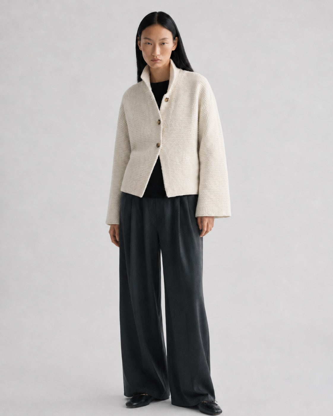 A model stands against a light gray studio background wearing a cropped, textured cream-colored jacket with brown buttons, a black top, and wide-leg charcoal gray trousers.