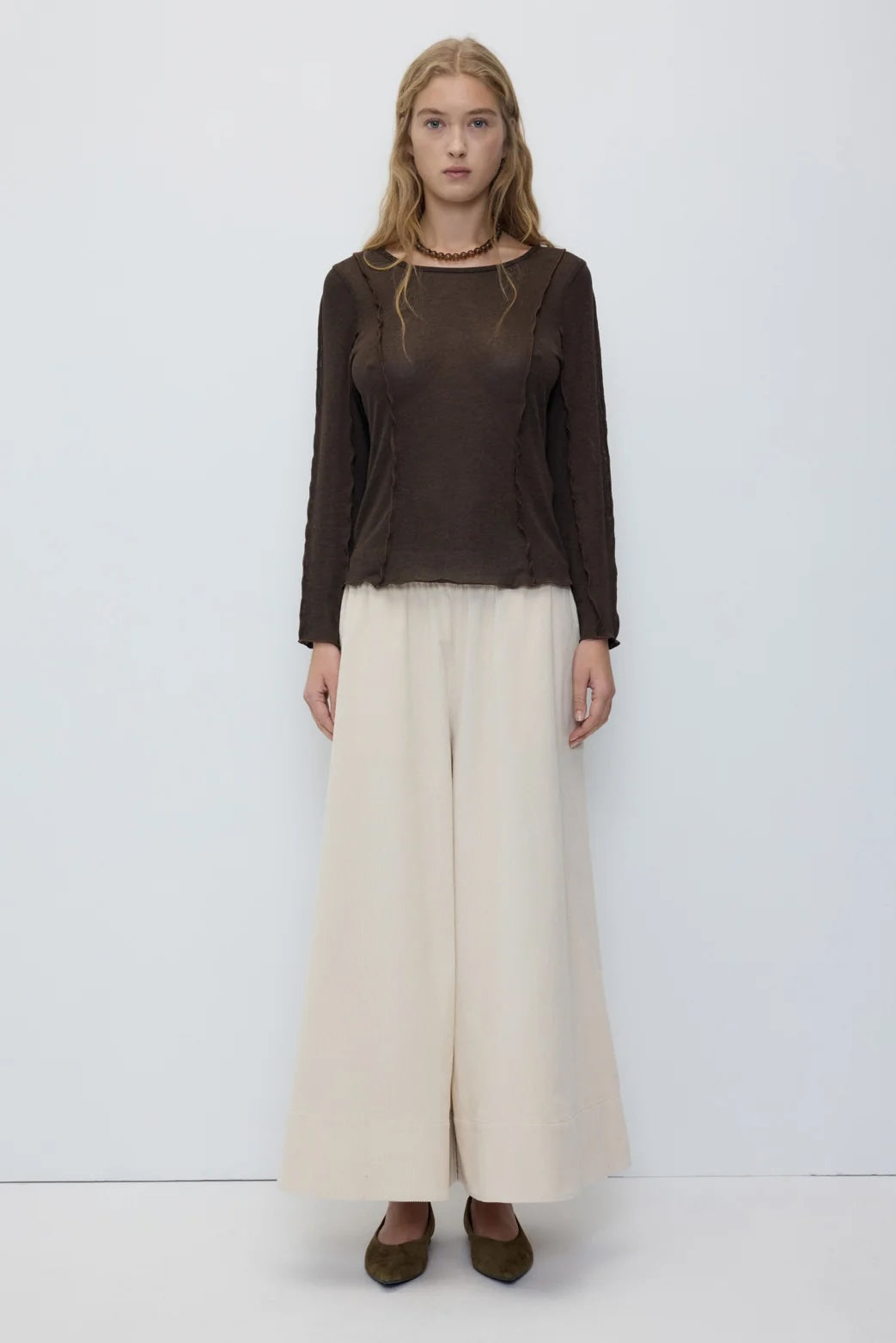 A woman stands against a plain light gray studio background wearing a dark brown long-sleeve sheer top and wide-leg cream pants.