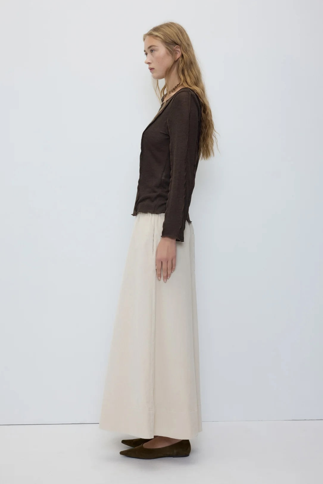 A woman stands in profile wearing a dark brown long-sleeve top and a long cream skirt against a plain light gray studio background.