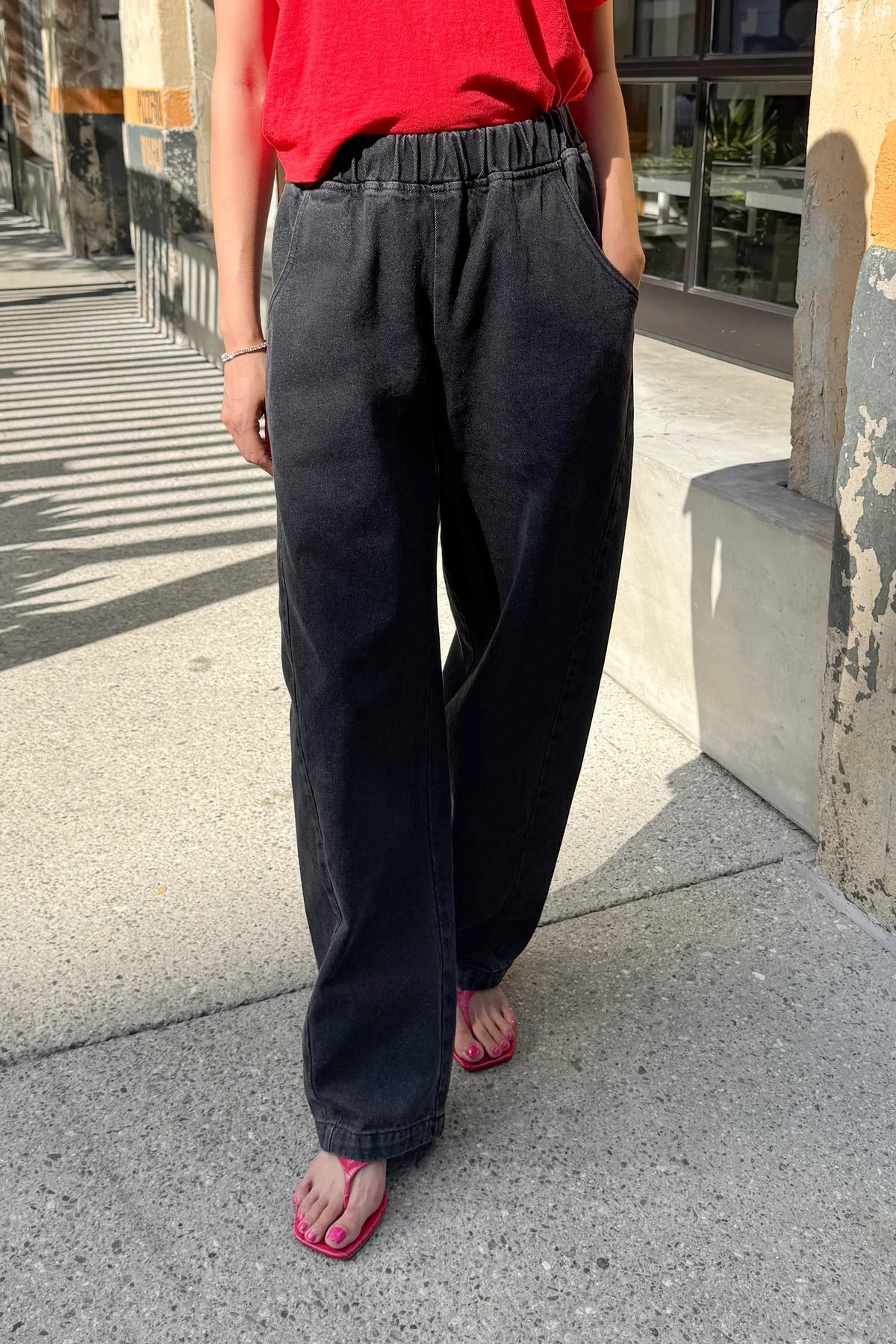 A person wearing loose, wide-leg dark charcoal gray jeans with an elastic waistband, standing on a concrete sidewalk outdoors.