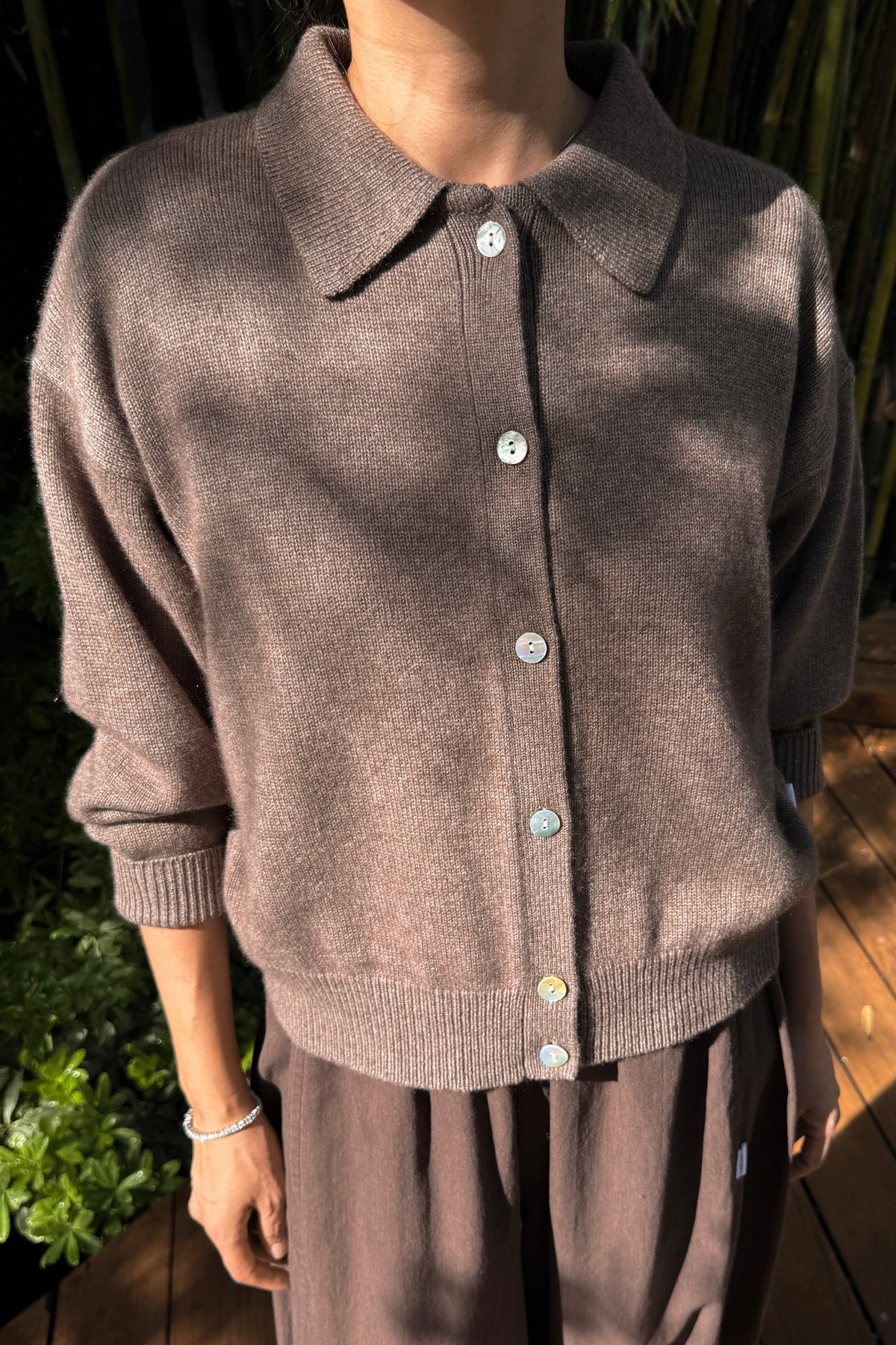 A person wearing a taupe-brown knit cardigan with a collar and front buttons, paired with matching brown pants, photographed outdoors with dappled sunlight and greenery in the background.