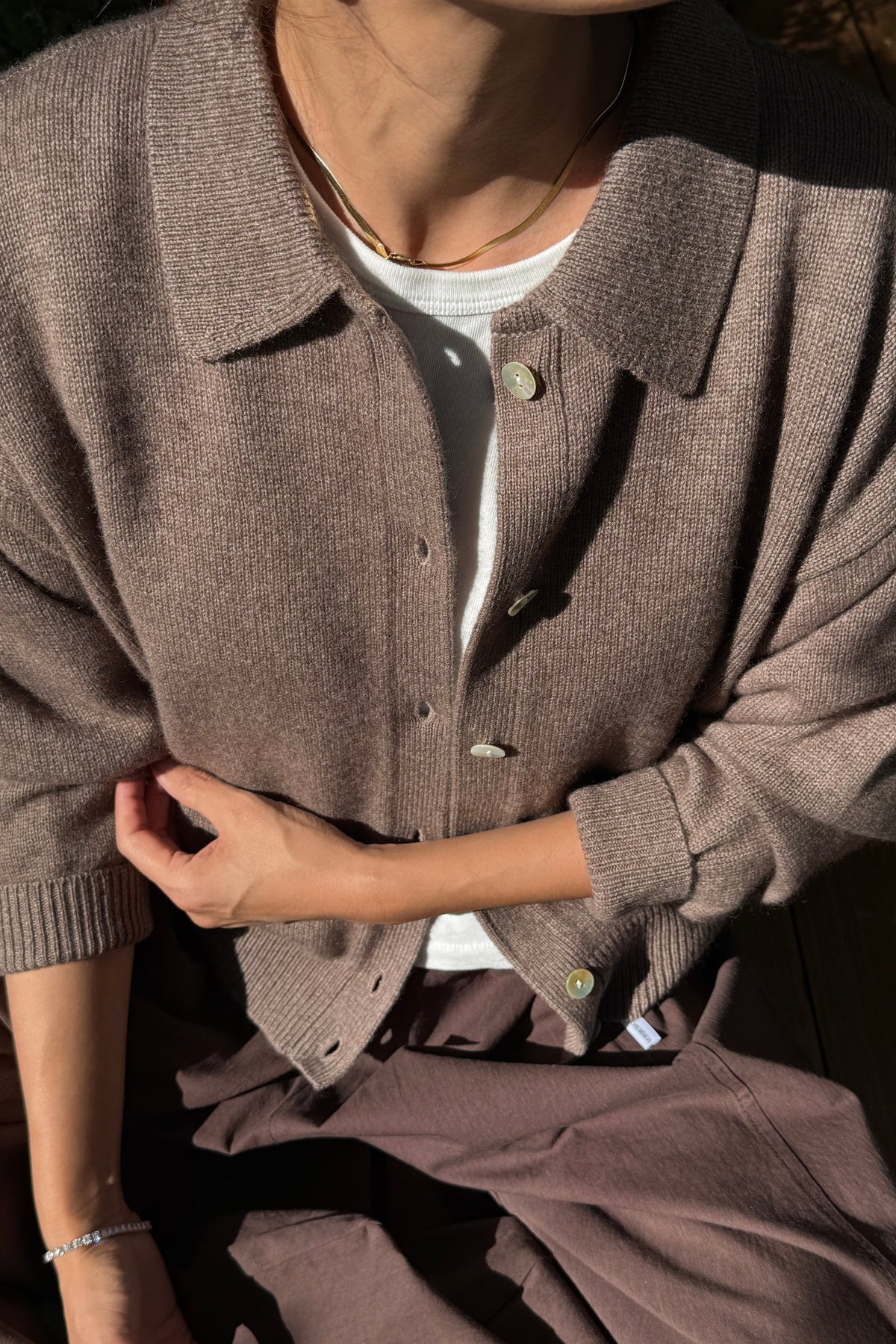 A person wearing a taupe-brown ribbed knit button-front cardigan over a white undershirt, paired with matching brown trousers. The photo is taken in warm natural light with strong shadows, focusing on the texture of the cardigan.
