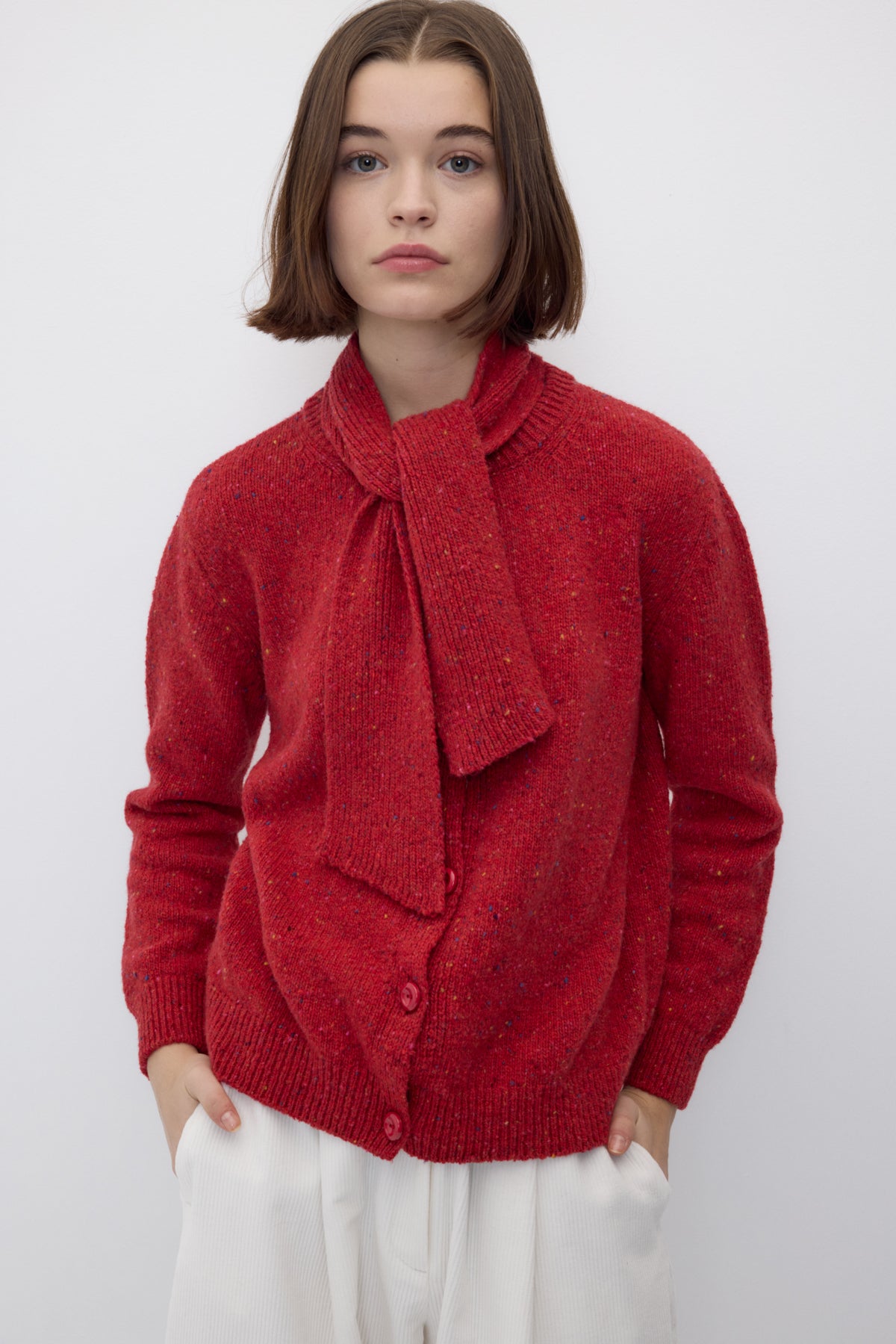 A model wearing a textured red knit cardigan with a large bow detail at the collar and white trousers, photographed against a neutral light gray background.