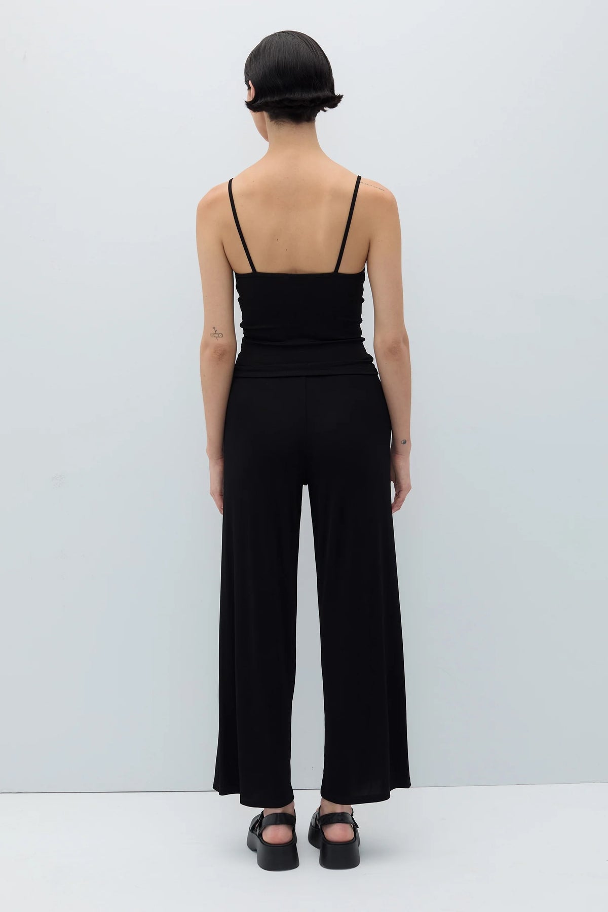 A model viewed from the back wearing a black spaghetti-strap camisole top and matching wide-leg trousers, paired with black platform sandals against a light gray studio background.