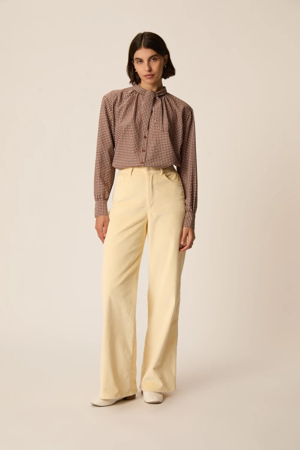 A model stands against a plain beige studio background wearing a brown and beige gingham long-sleeve blouse with a high neck and tie collar, paired with cream wide-leg trousers.