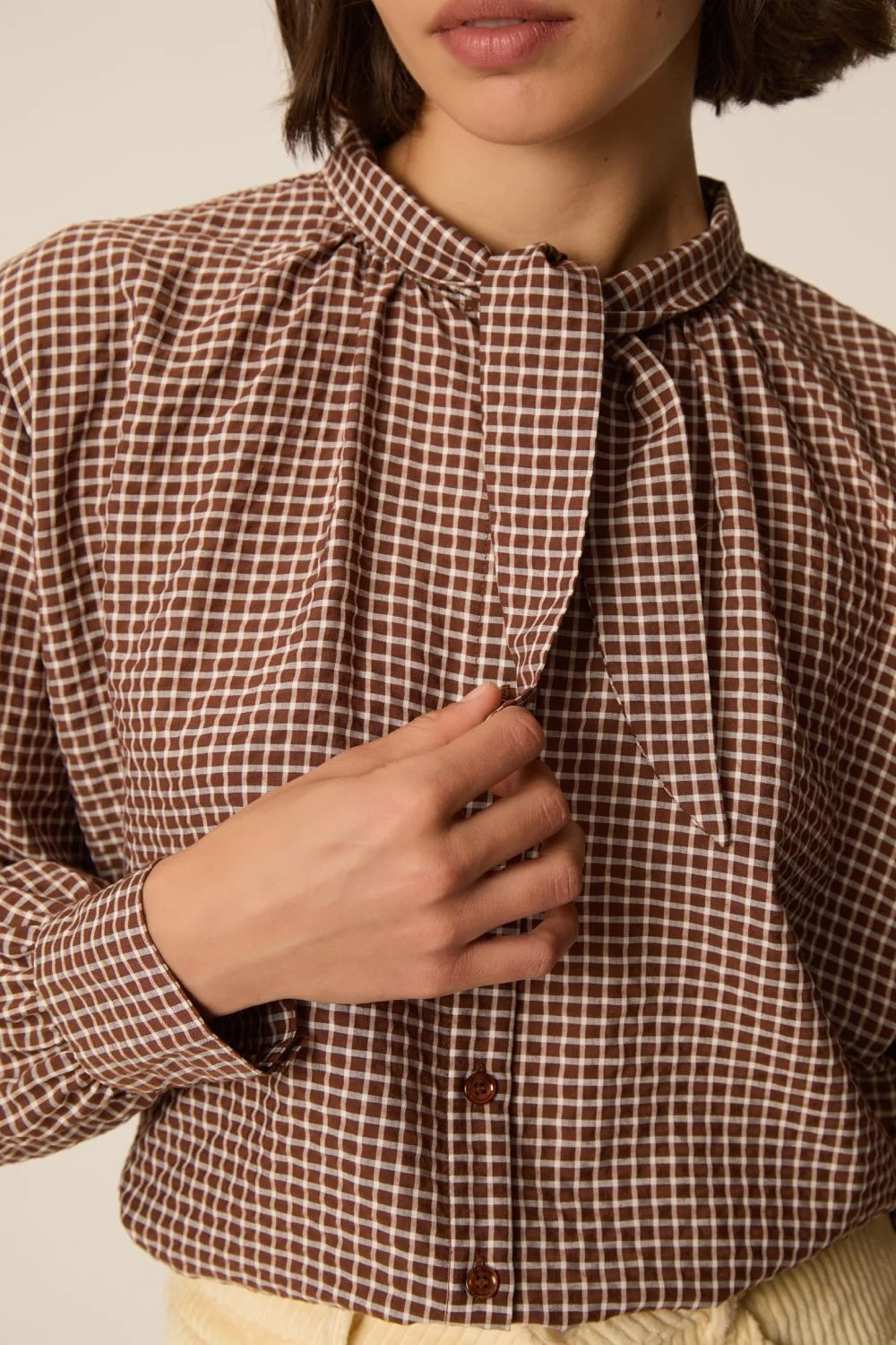 A close-up of a person wearing a brown and beige gingham check long-sleeve blouse with a high gathered collar and front buttons.