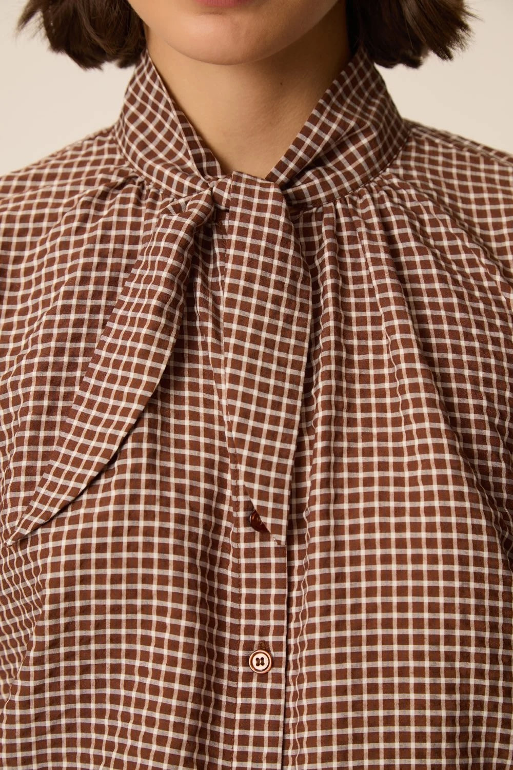 Close-up of a person wearing a brown and beige gingham blouse with a large bow tie collar and front button closure.
