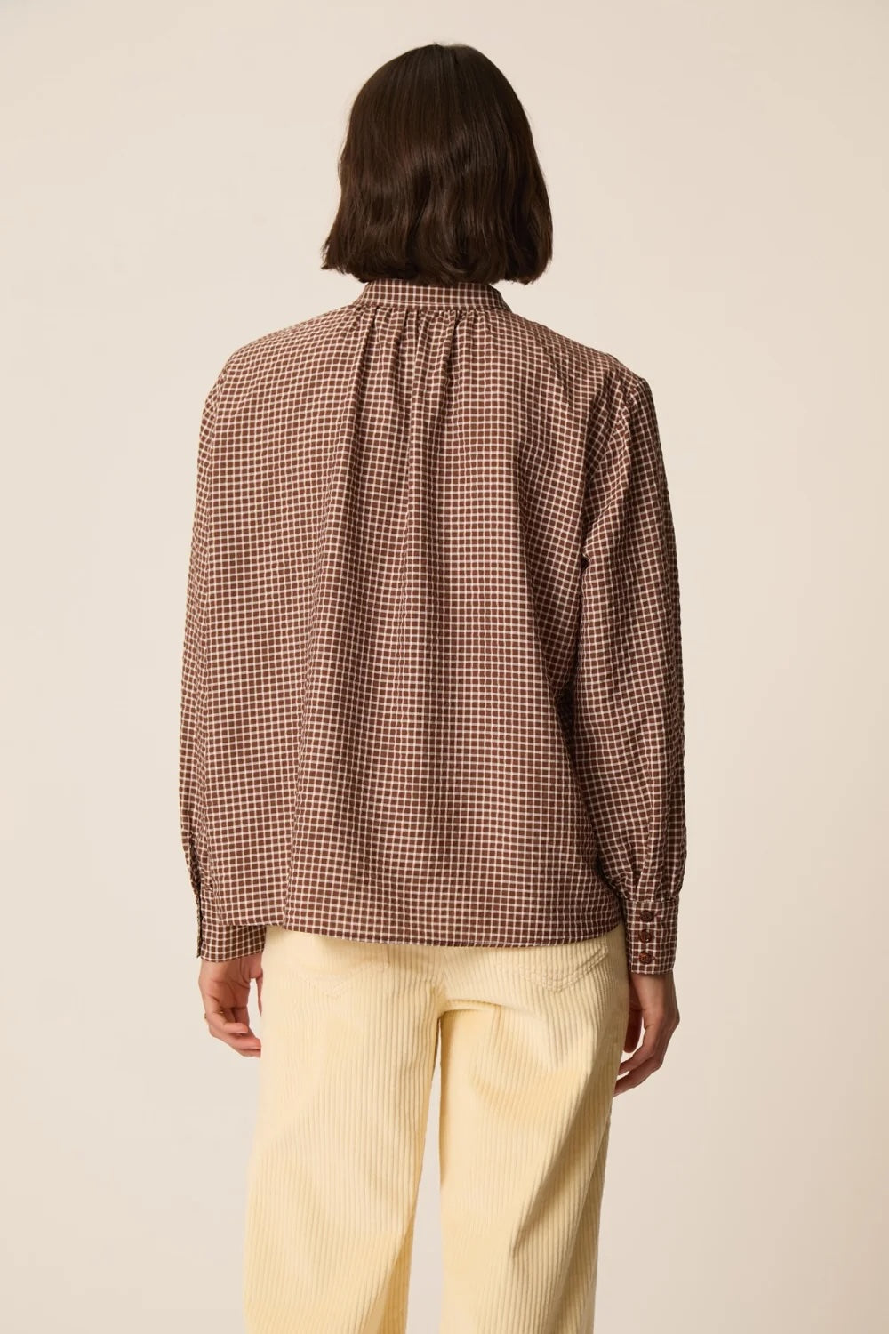 A model shown from the back wearing a brown and beige checkered long-sleeve blouse with a gathered neck, paired with light yellow trousers, against a plain beige background.