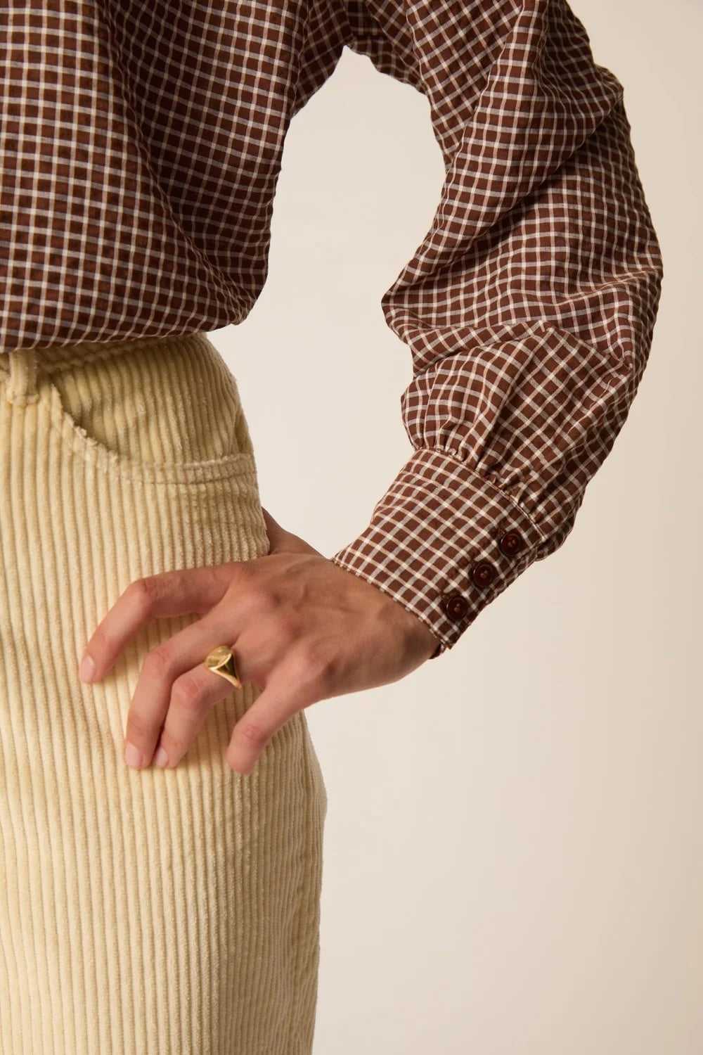 A close-up of a person wearing a brown and beige gingham long-sleeve shirt and a cream corduroy skirt, with one hand resting on the hip. A gold ring is visible on the finger, set against a plain beige background.