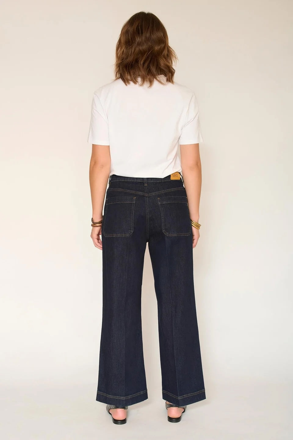 A woman viewed from behind wearing dark blue wide-leg denim jeans, a white t-shirt, and black sandals against a plain off-white background.
