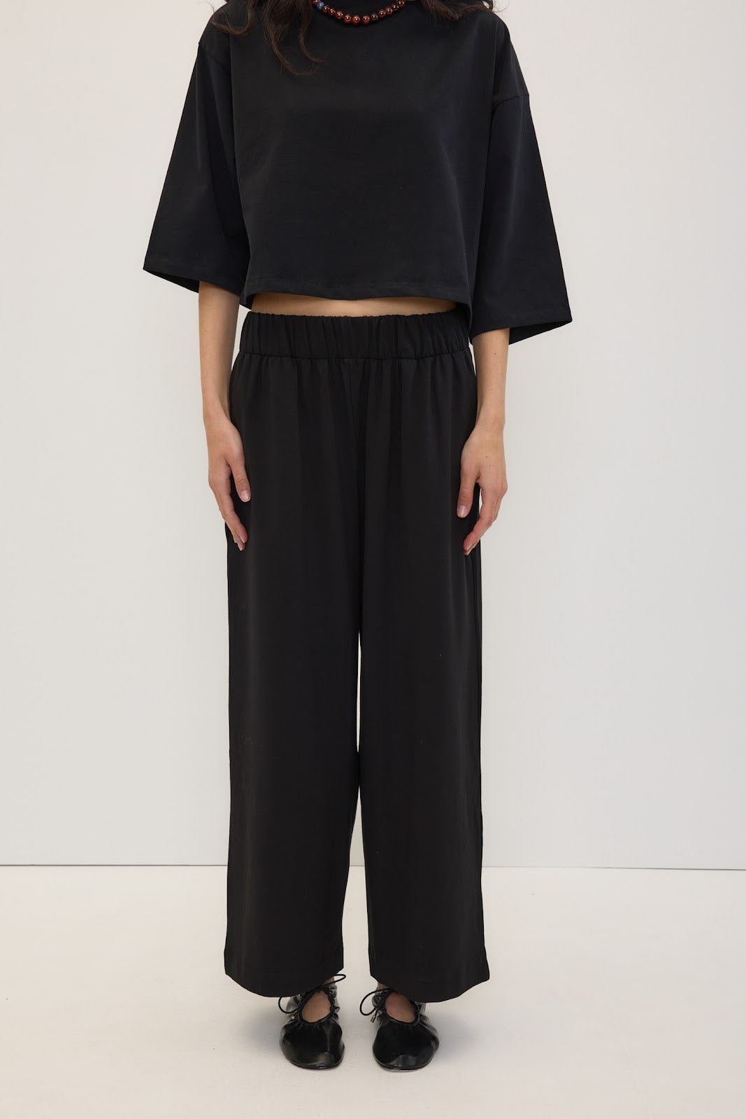 A model wearing a black short-sleeve cropped t-shirt and matching black wide-leg trousers, paired with black shoes and a beaded necklace, standing against a plain white studio background.