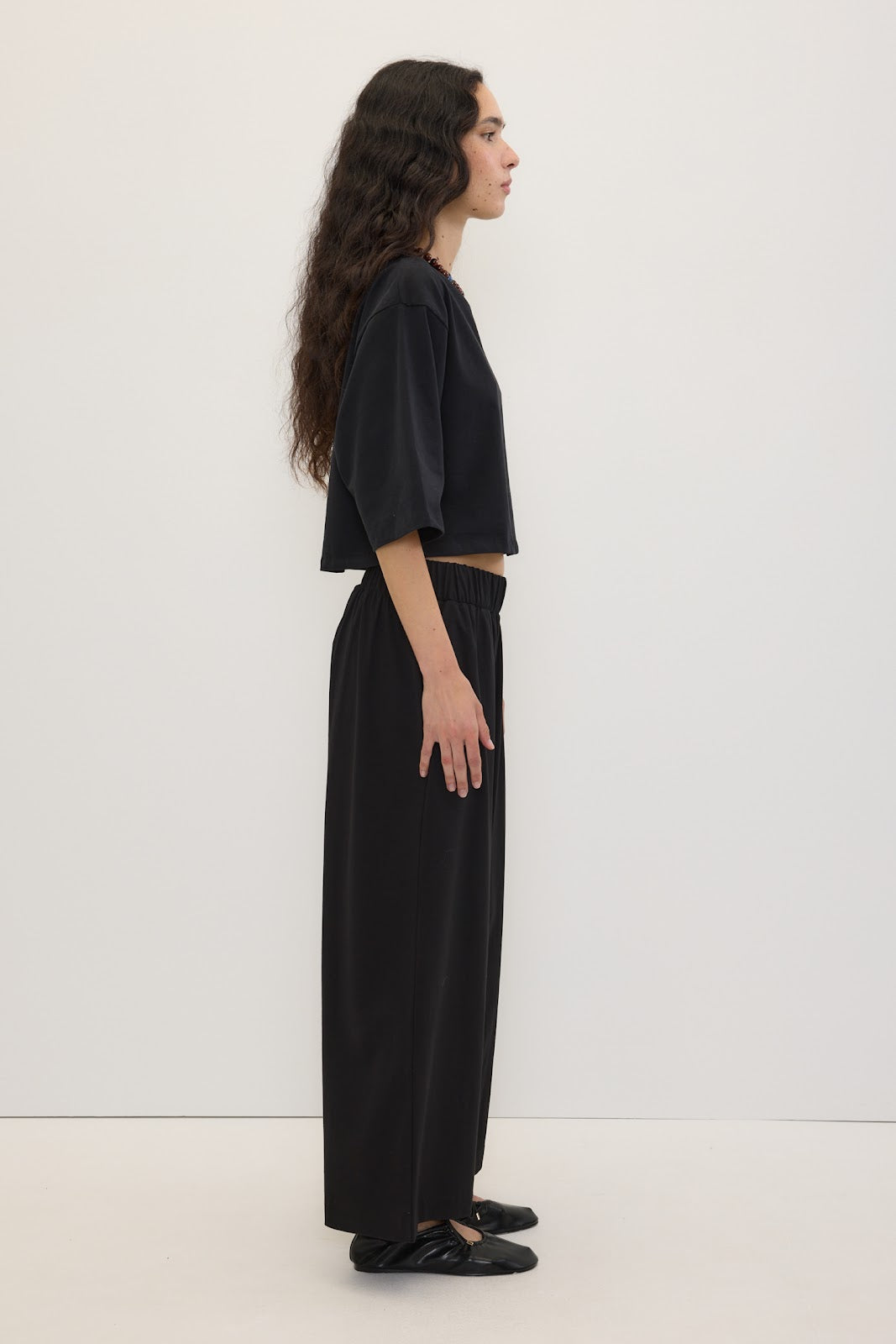 A woman stands in profile wearing a black cropped short-sleeve top and black wide-leg pants against a plain white studio background.
