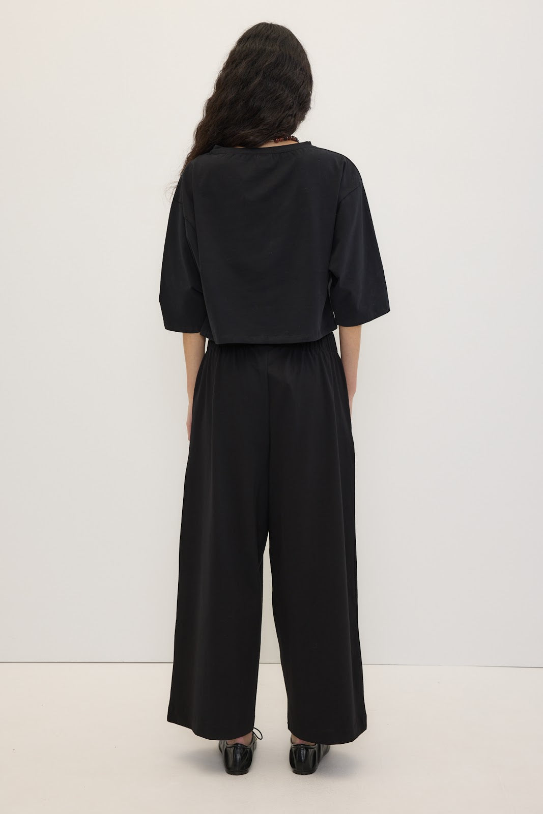 A model viewed from the back wearing a black short-sleeve top and matching wide-leg pants, standing against a plain white studio background.