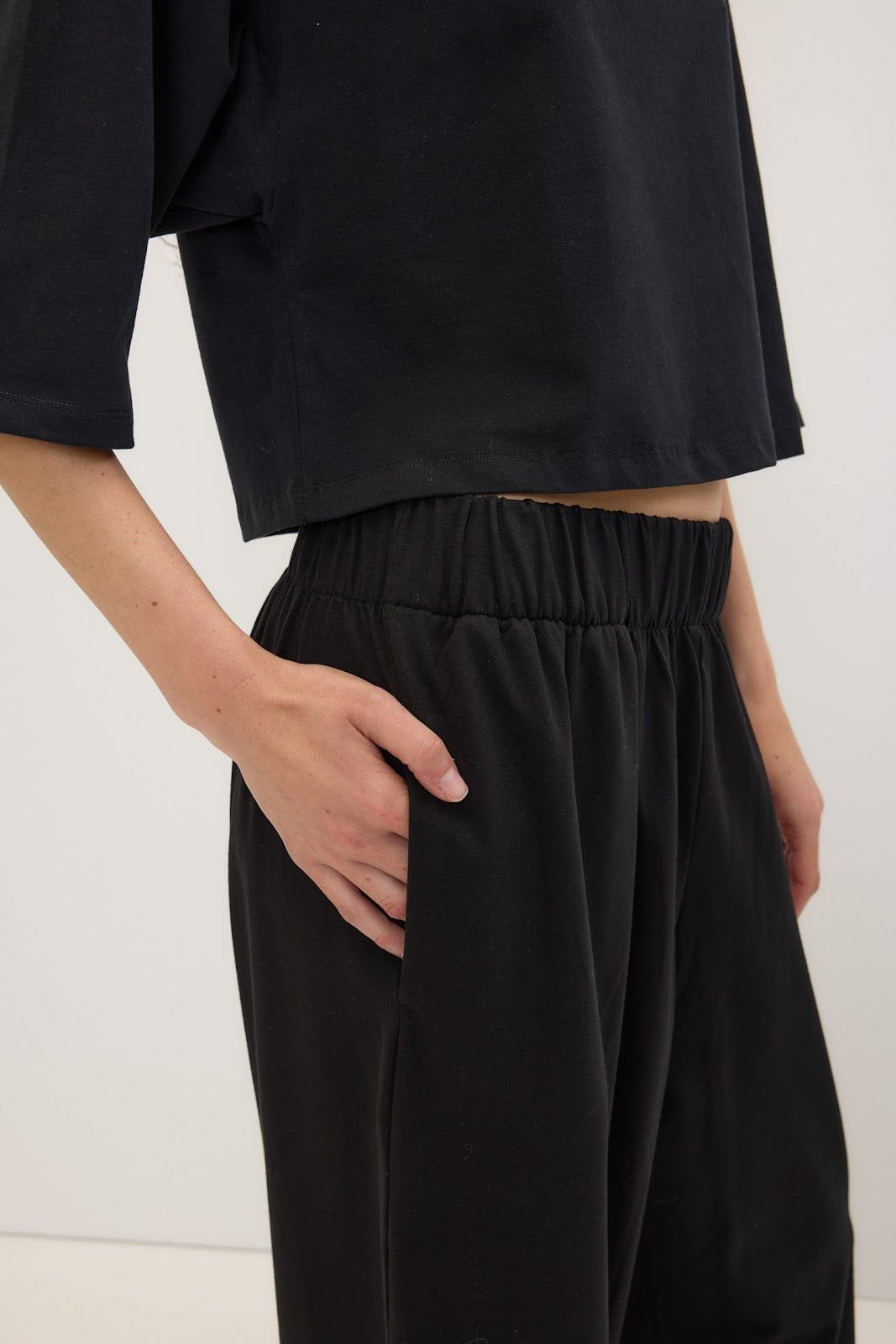 A close-up of a person wearing black wide-leg trousers with an elastic waistband and a black cropped top, standing against a plain light background.