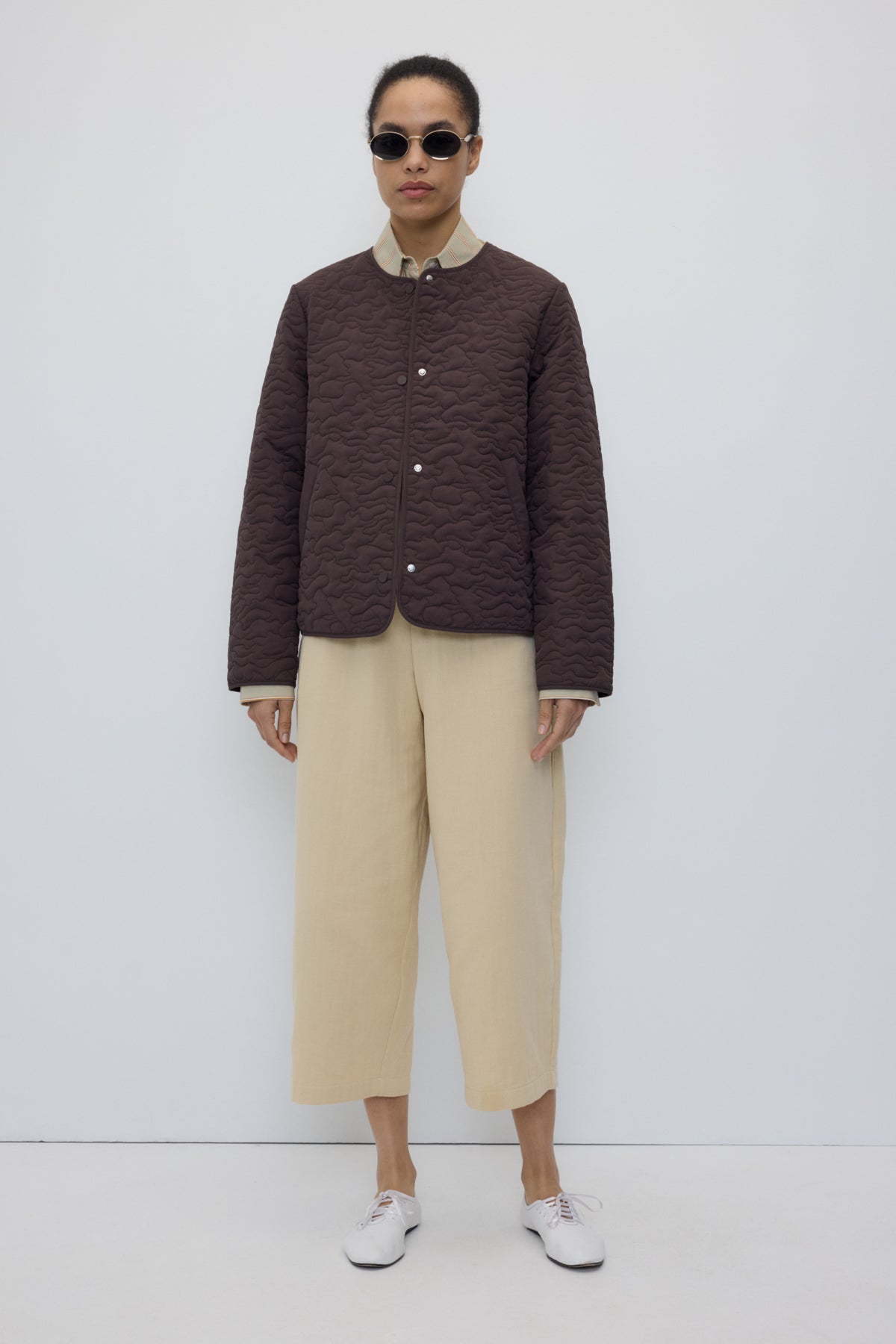 A model stands against a plain white studio background wearing a dark brown textured quilted jacket with a light beige collar and cuffs, beige cropped wide-leg trousers, and white sneakers.