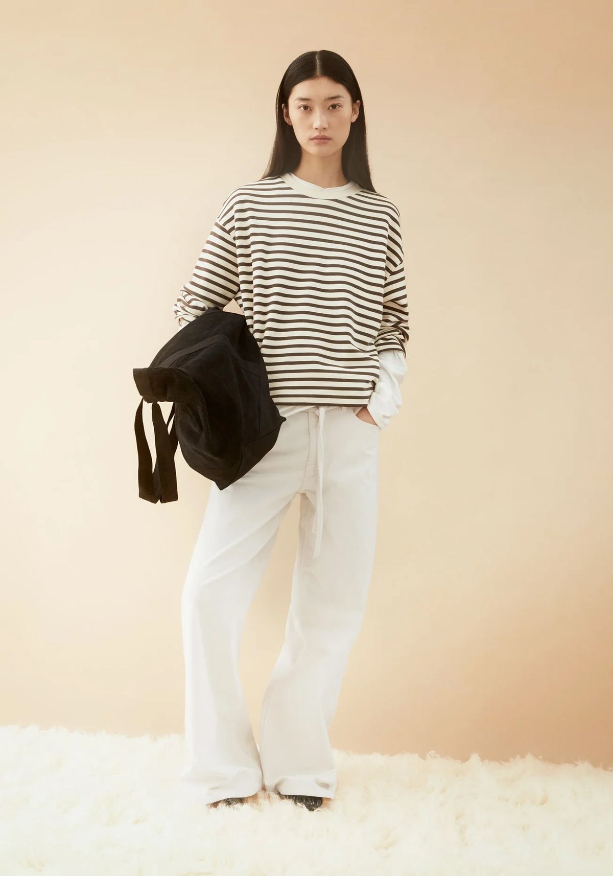 A model stands against a warm beige studio backdrop wearing a black and white horizontally striped long-sleeve top, white wide-leg pants, and holding a black backpack.