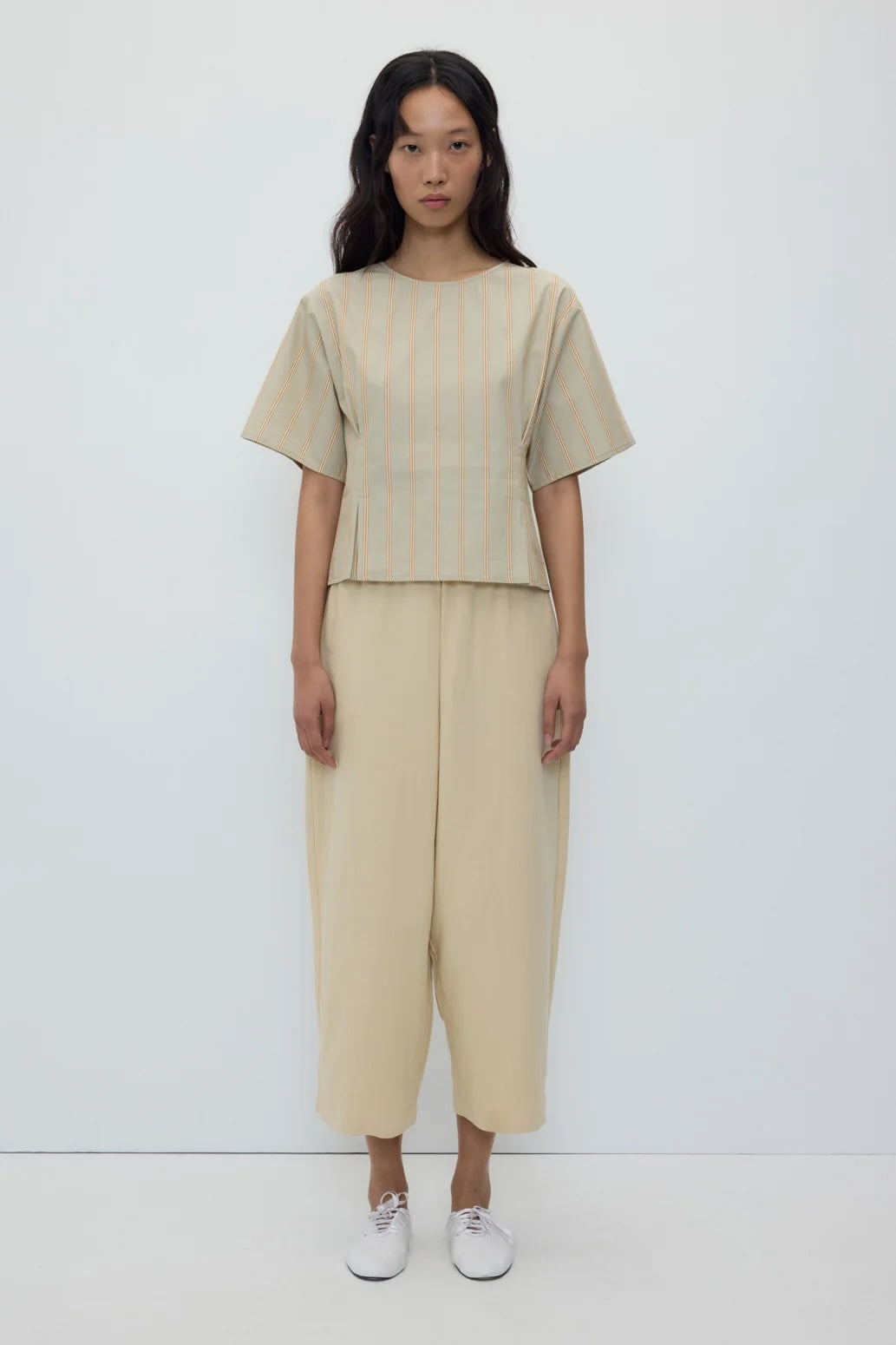 A model stands against a plain white studio background wearing a beige short-sleeve top with subtle vertical stripes and matching wide-leg cropped pants, paired with white sneakers.