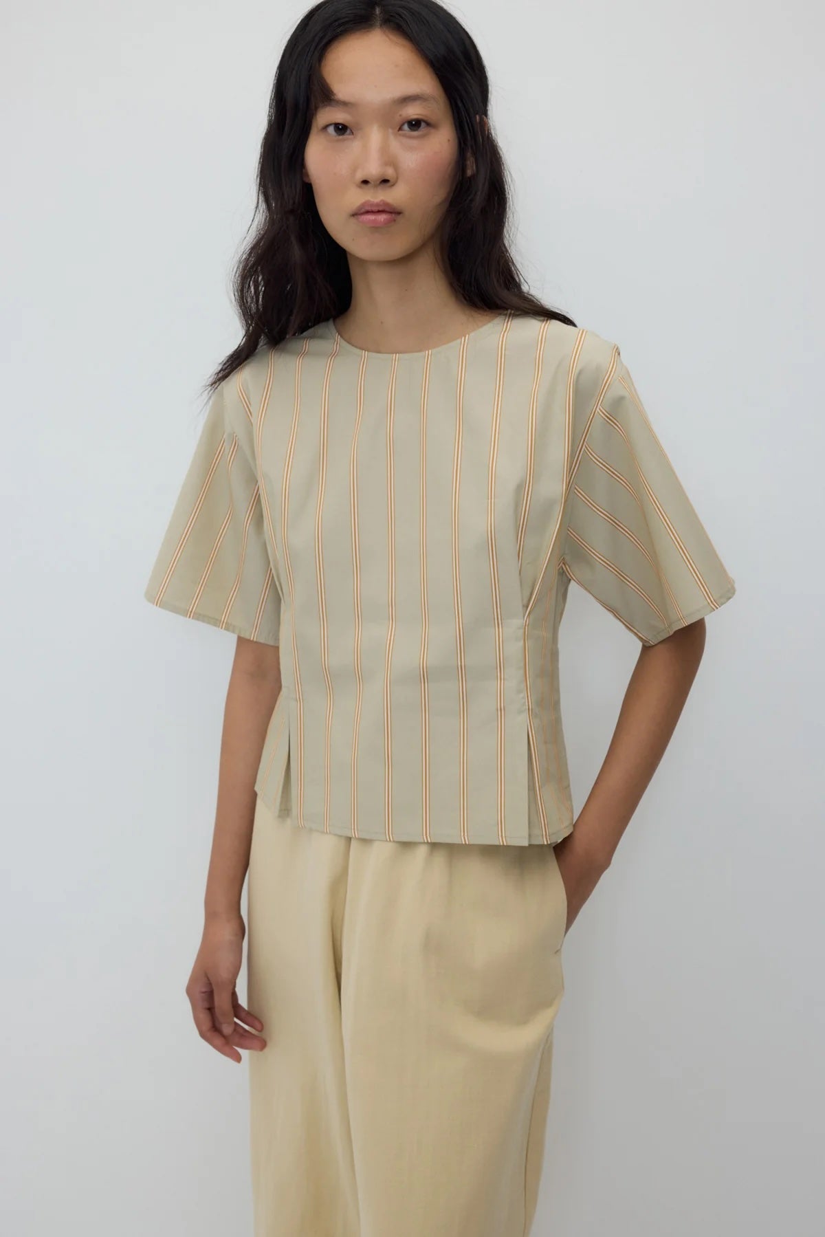 A model wearing a beige short-sleeve top with thin orange vertical stripes and matching beige wide-leg trousers, standing against a plain light gray background.