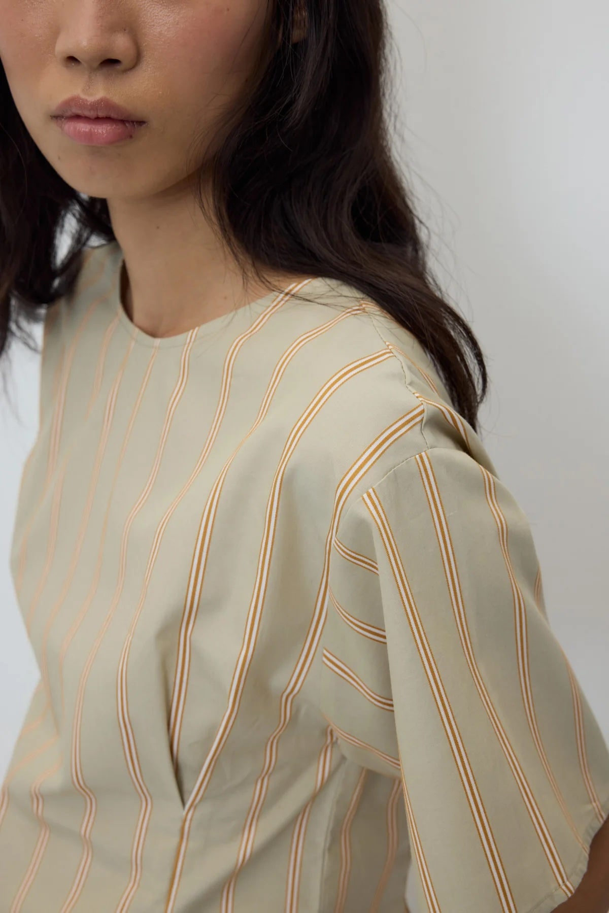 Close-up of a model wearing a short-sleeve top in a light beige or cream color with thin vertical peach-orange stripes, set against a plain light gray background.
