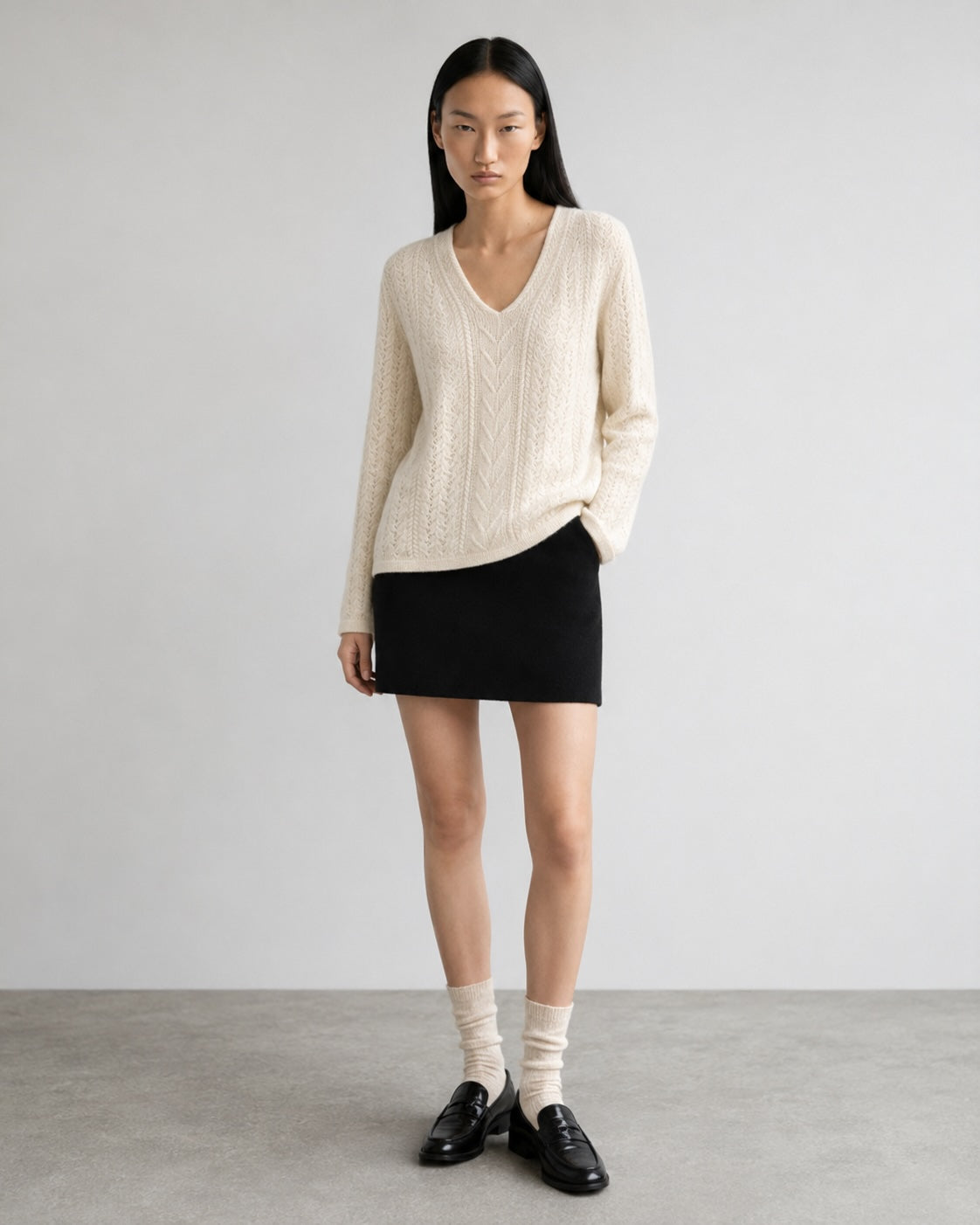A model stands in a studio wearing a cream cable-knit V-neck sweater, a black mini skirt, cream socks, and black leather loafers against a light gray background.