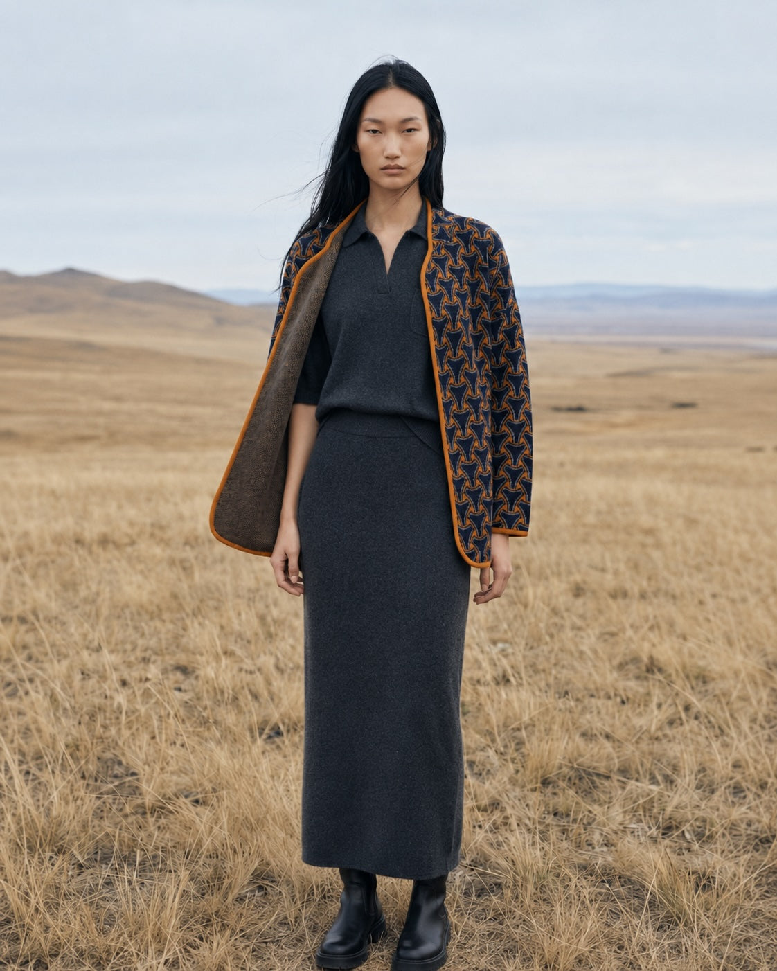 A model stands in an open grassland wearing a dark charcoal gray knit long-sleeve top and matching skirt, styled with a patterned brown and navy quilted jacket featuring orange-brown trim.