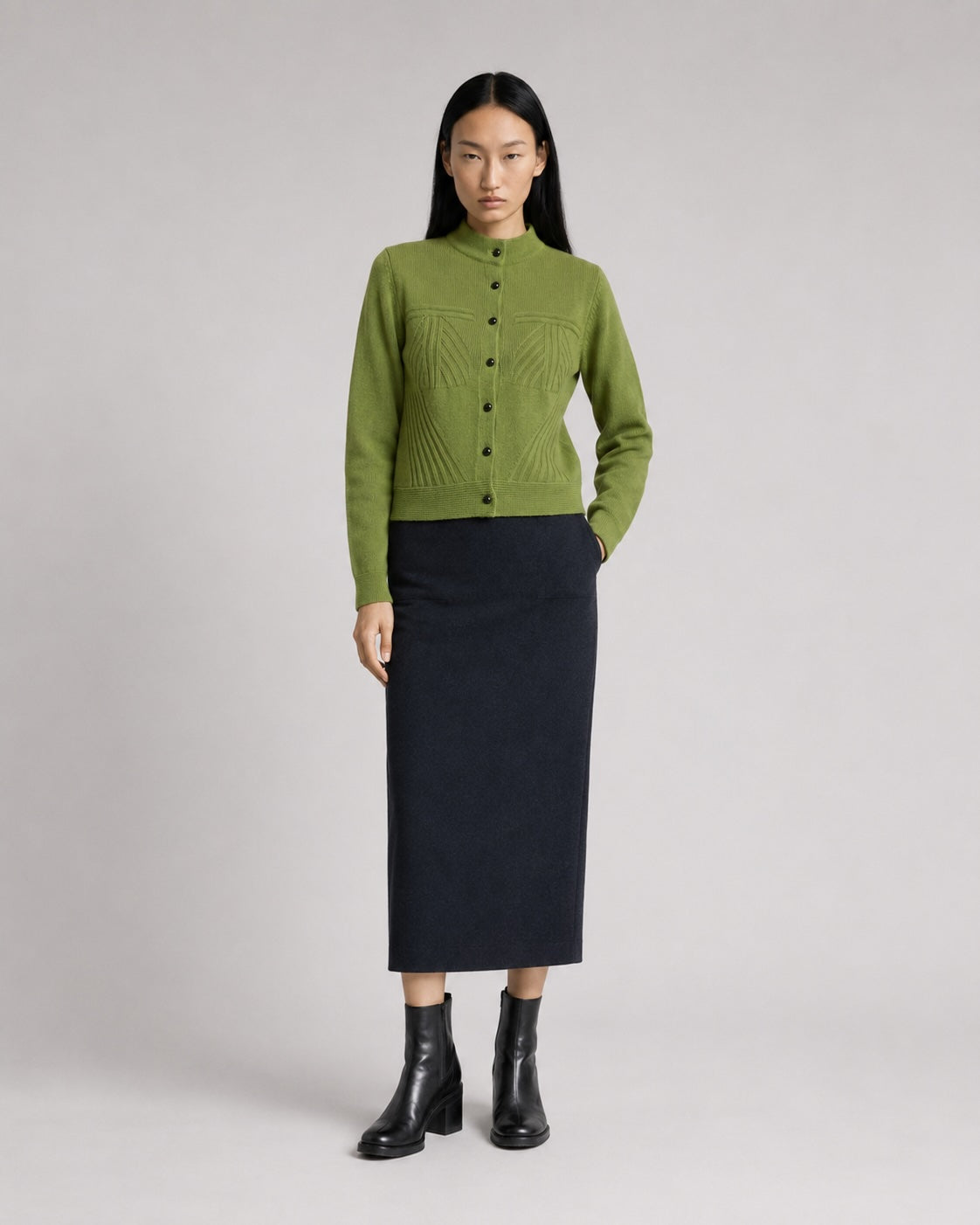 A model stands against a light gray studio background wearing a green button-front knit cardigan, a dark navy midi skirt, and black ankle boots.