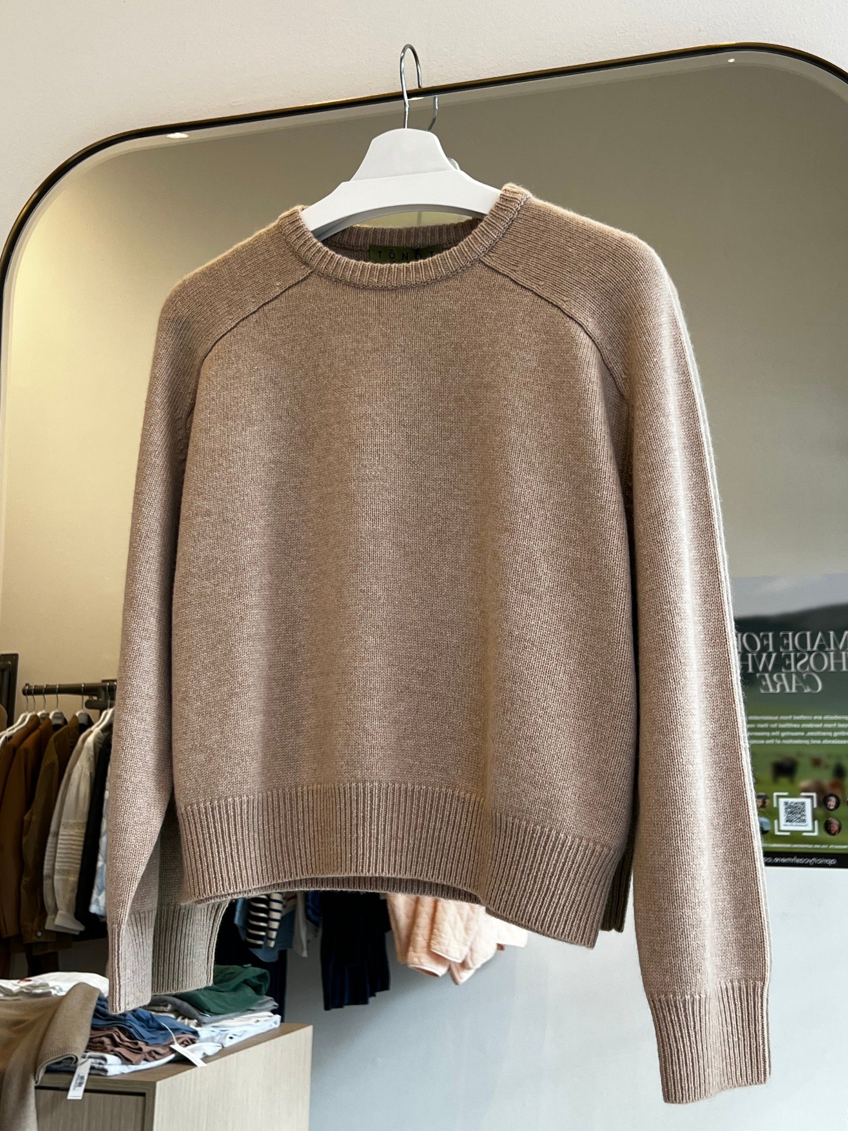 Beige round neck cropped cashmere sweater with raglan sleeves hanging on white hanger in store setting