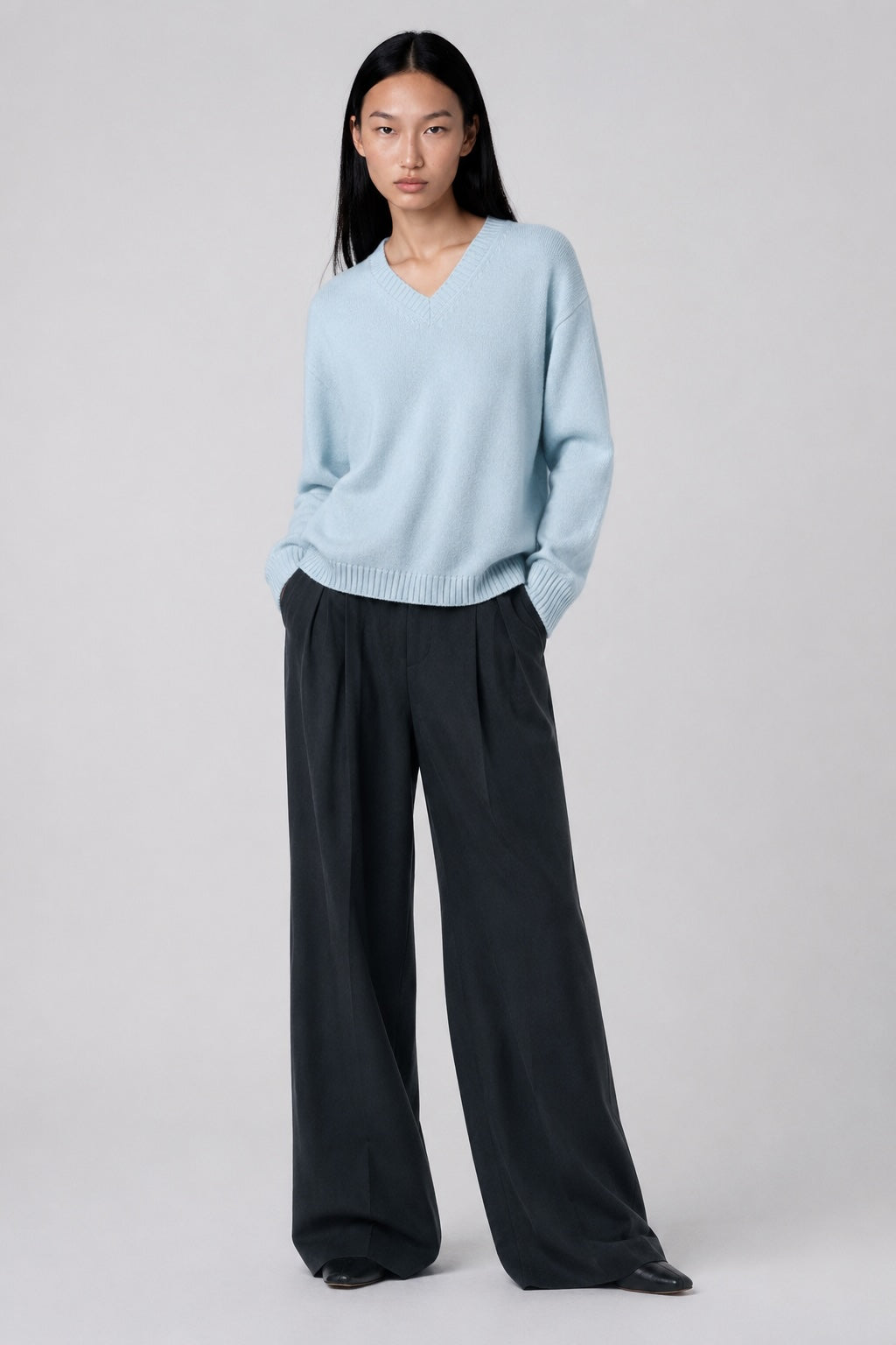 A model stands against a light gray studio background wearing a light blue v-neck knit sweater and dark charcoal wide-leg trousers.
