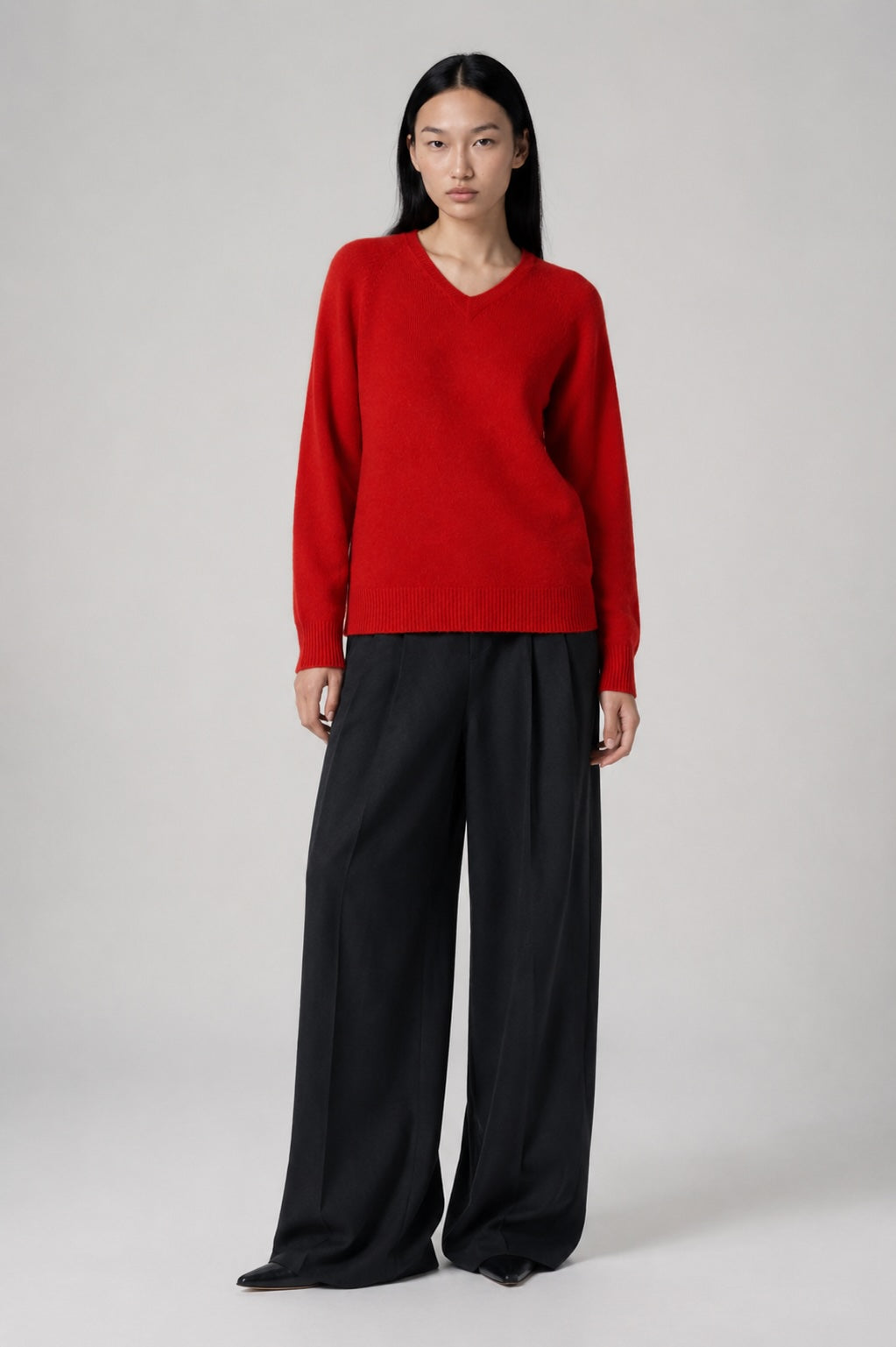A model stands against a light gray studio background wearing a bright red long-sleeve V-neck sweater and black wide-leg trousers.