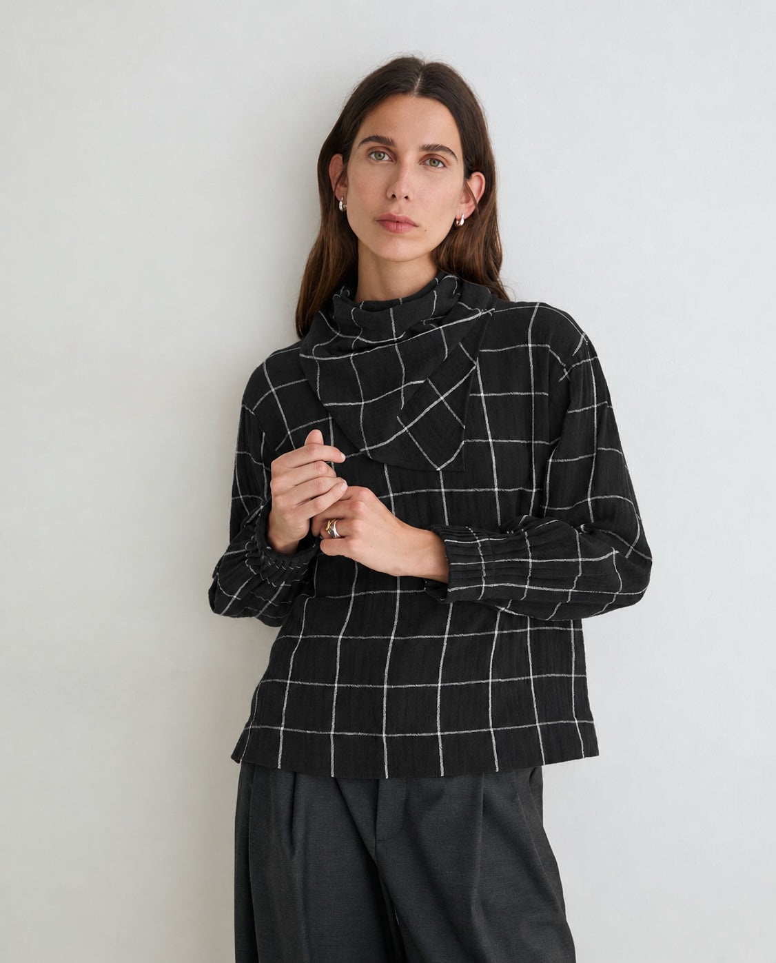 A woman wearing a black long-sleeve top with a white windowpane grid pattern and dark gray wide-leg trousers, standing against a light gray wall.