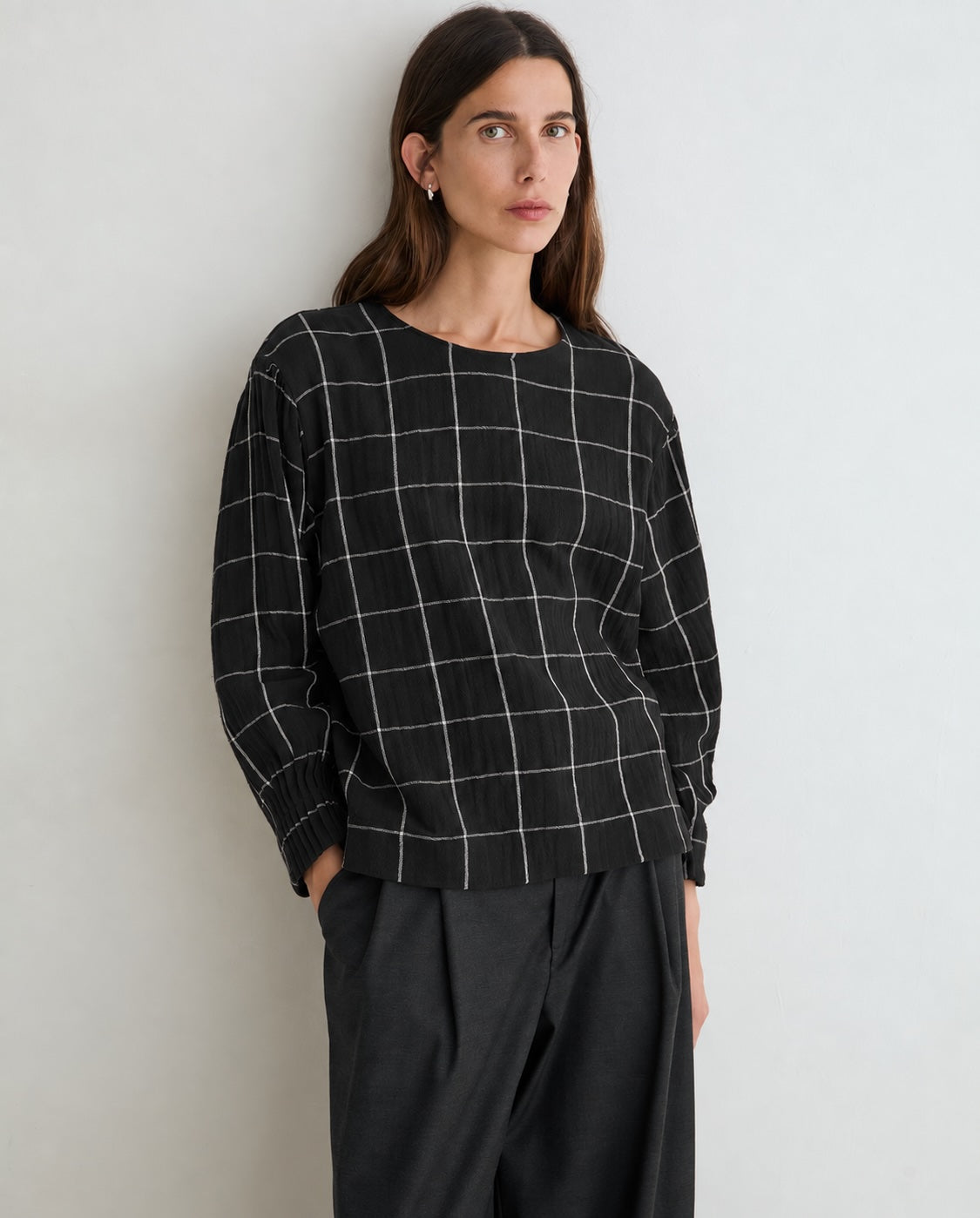 A woman wearing a black long-sleeve top with a white windowpane grid pattern and black wide-leg trousers, standing against a plain light gray wall.