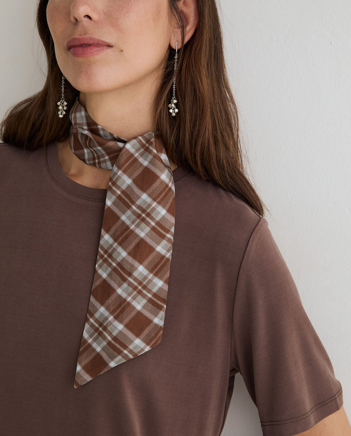 A close-up of a woman wearing a brown and beige plaid scarf tied around her neck, paired with a brown crewneck t-shirt and dangling earrings.