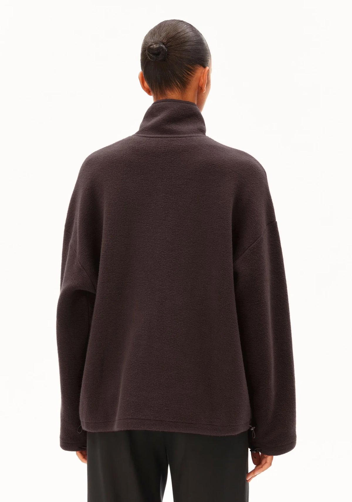 A person shown from the back wearing an oversized dark brown mock-neck or turtleneck top and black pants against a clean white studio background.