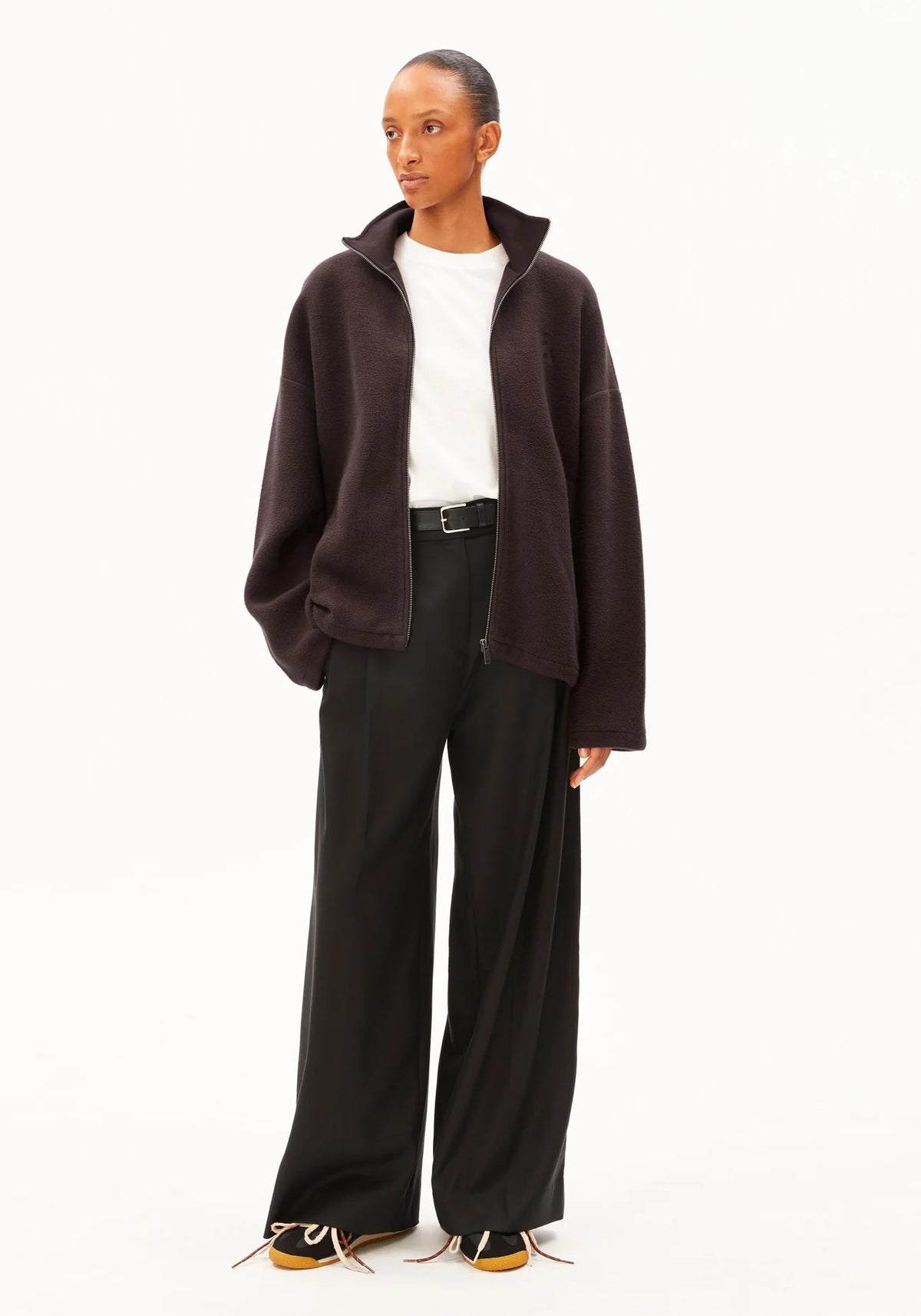 A model stands against a plain white studio background wearing a dark brown fuzzy zip-up jacket over a white t-shirt, black wide-leg trousers, and brown sneakers.