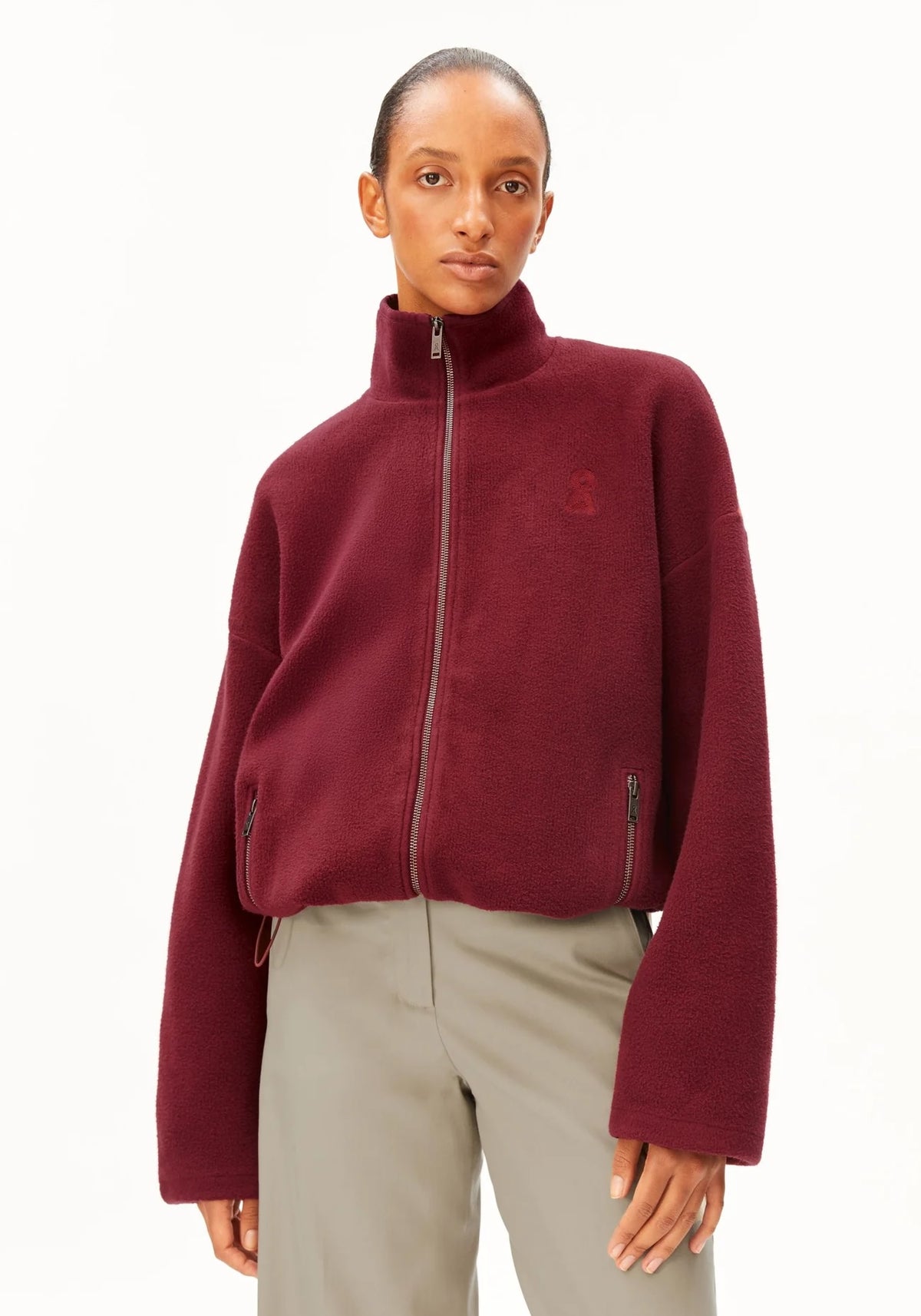A model wearing a cropped, long-sleeve fleece jacket in deep burgundy with a high collar and a subtle tonal embroidered logo on the left chest, paired with light gray trousers, against a plain white background.