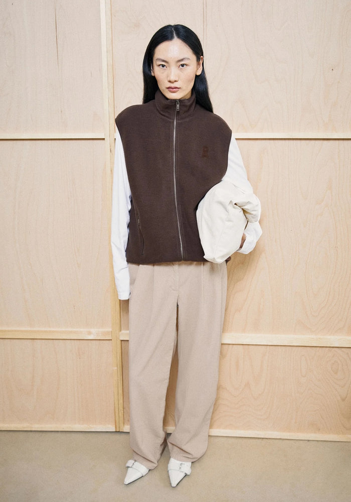 A model stands against a light wood-paneled wall wearing a dark brown zip-up fleece vest over a white long-sleeve top, paired with beige wide-leg trousers and white pointed shoes, holding a white shoulder bag.