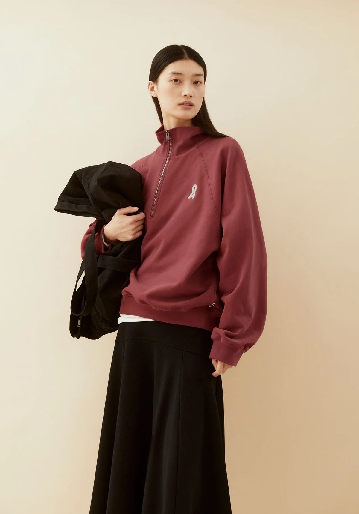 A model stands against a plain beige background wearing a dusty rose quarter-zip sweatshirt with a small white ribbon graphic on the chest, paired with a long black skirt and holding a black jacket.