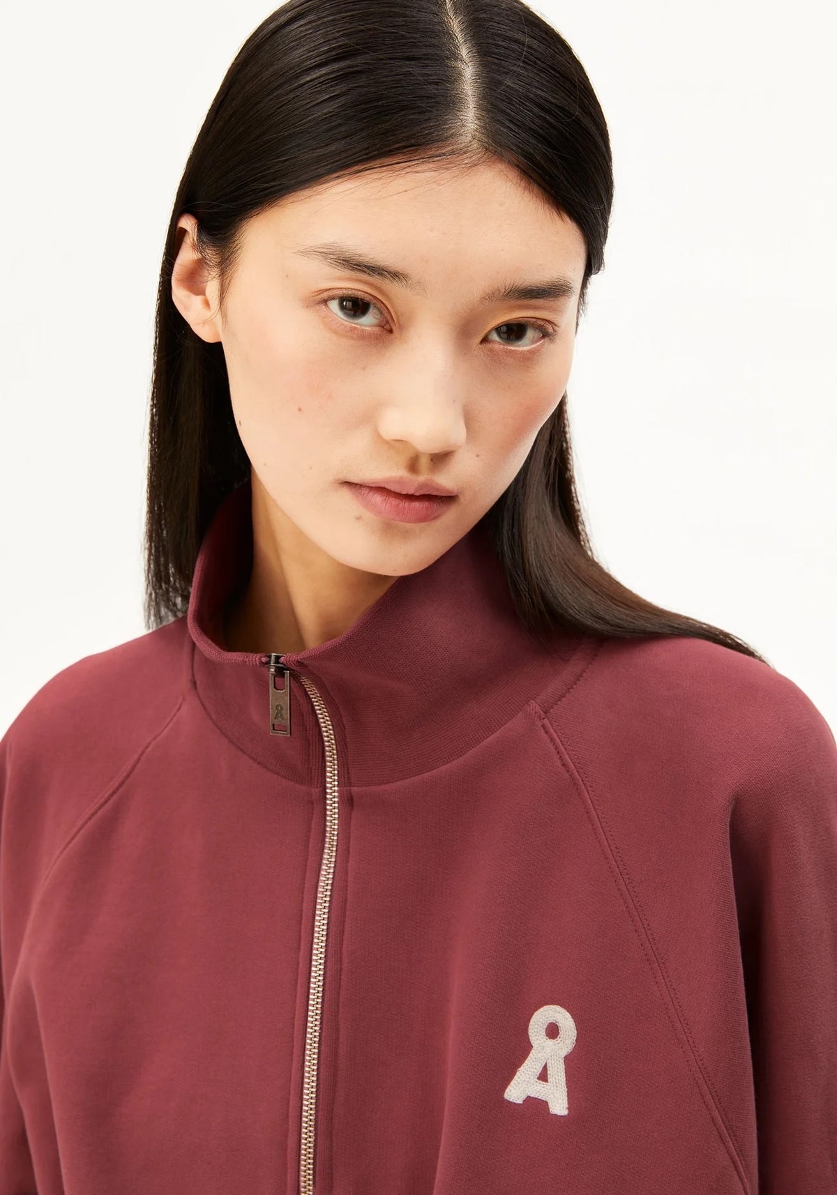 A close-up of a model wearing a burgundy zip-up sweatshirt with a small white logo on the left chest, set against a plain white background.