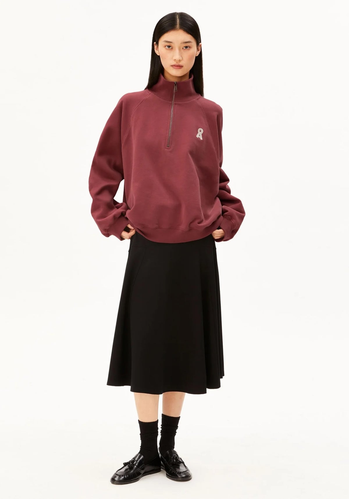 A model stands against a plain white studio background wearing a burgundy half-zip sweatshirt with a small white graphic on the left chest, a black midi skirt, black socks, and black leather shoes.