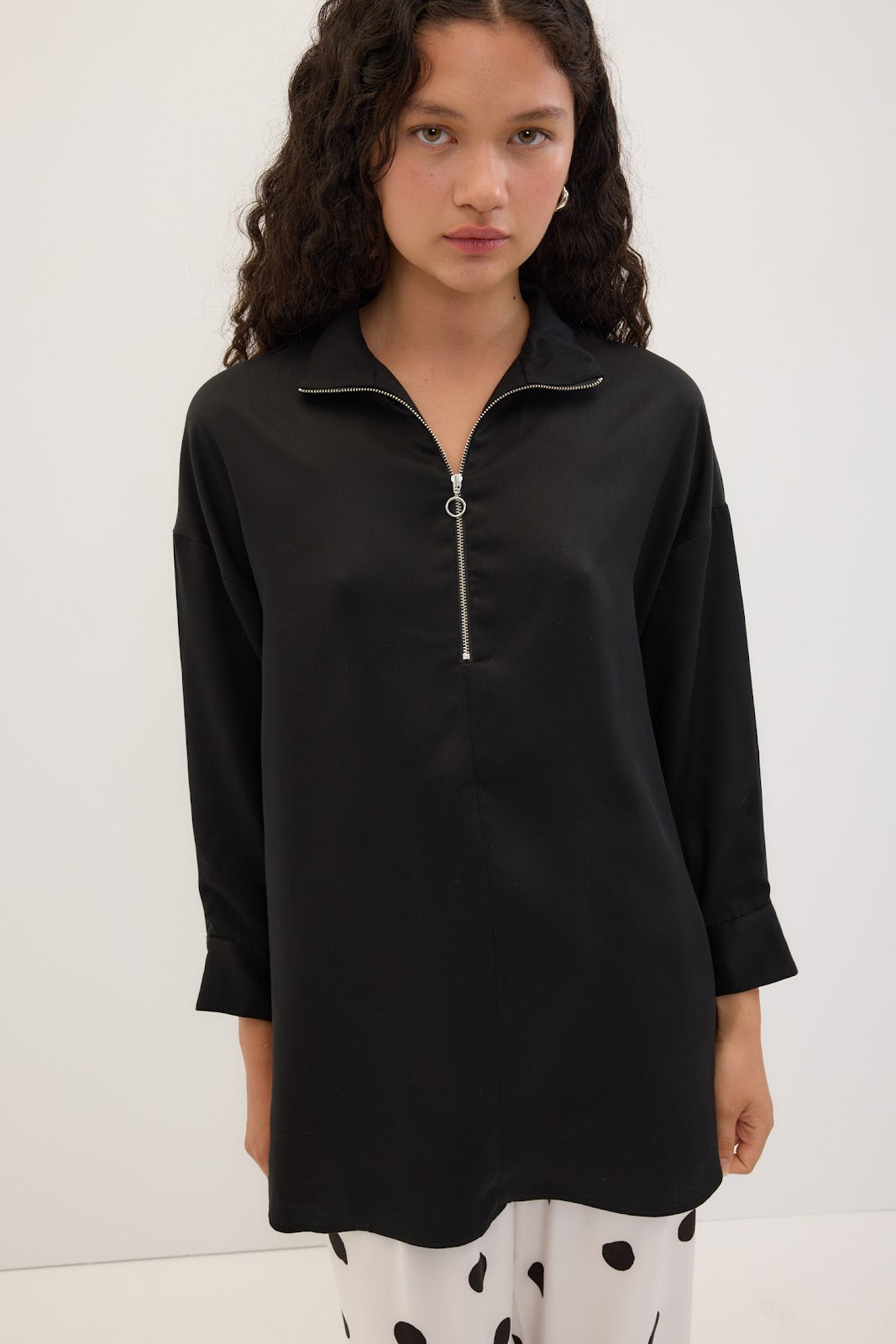 A model wearing a black long-sleeve tunic top with a half-zip collar, paired with white pants featuring black spots, set against a plain white studio background.