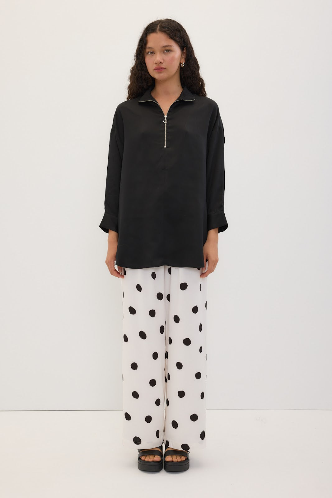 A model stands against a plain white studio background wearing a black long-sleeve half-zip top, white wide-leg pants with black polka dots, and black sandals.