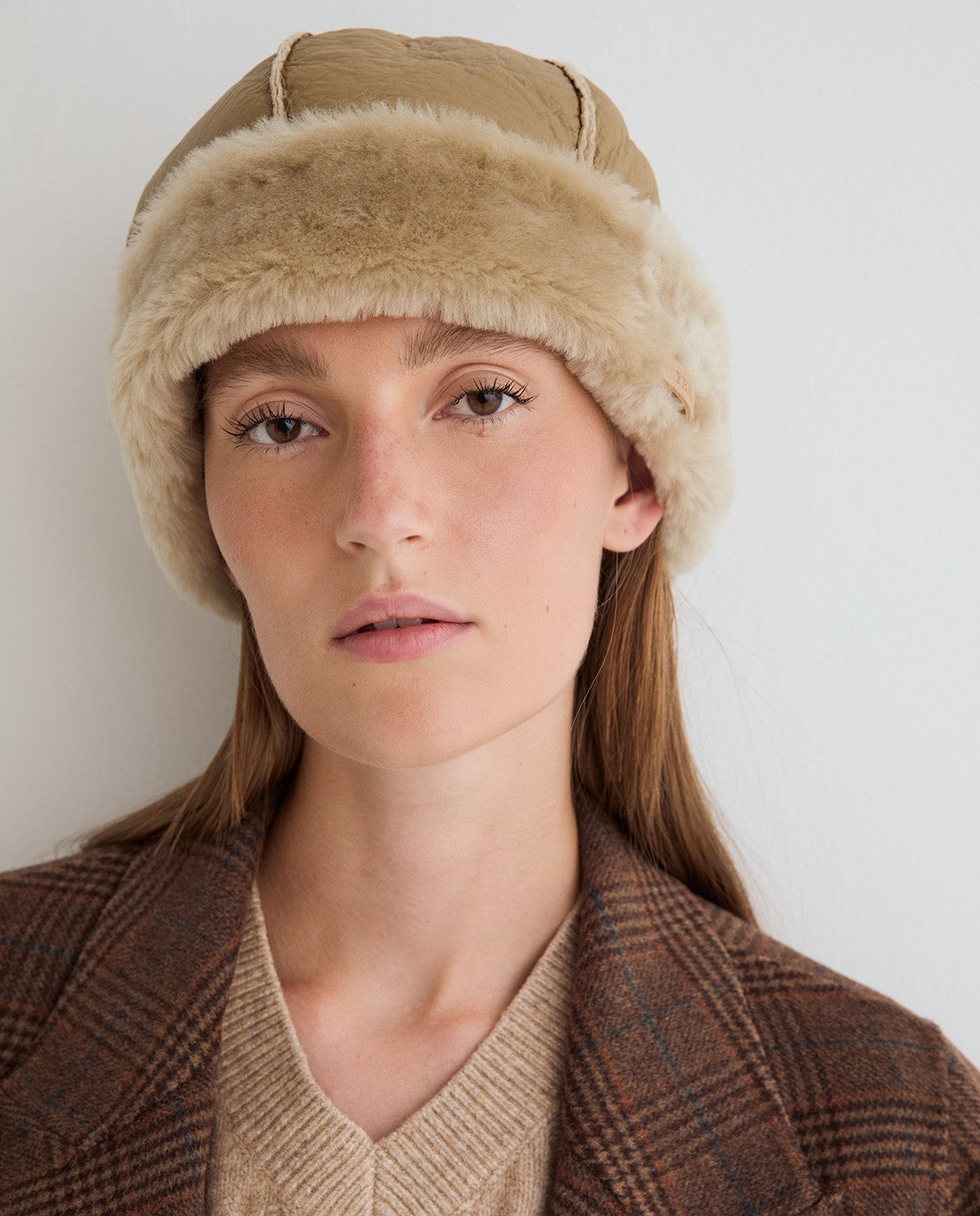 A close-up of a woman wearing a beige and brown fur-trimmed cap against a plain light background.